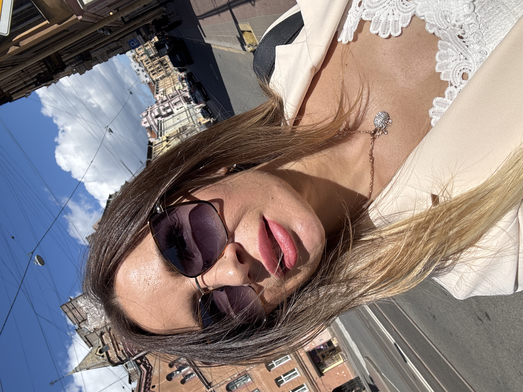 This is a close-up, self-portrait of a woman outdoors in Saint Petersburg, Russia. The camera angle is low, looking up towards the woman's face and shoulders. She is wearing sunglasses with dark, reflective lenses and a light-colored blazer over a white lace top. A delicate necklace with a jeweled pendant rests on her décolletage. Her lips are painted a rosy pink and are slightly parted. Her hair is long and appears to be a mix of brown and blonde, with strands falling over her face and shoulders.
The background is a blurred cityscape under a bright blue sky with scattered white clouds. The reflection in her sunglasses shows buildings and the sky. The image is taken during the daytime, likely on a sunny day, given the strong shadows and bright light. The architecture visible in the reflection suggests an urban environment, consistent with Saint Petersburg. There is no discernible text in the image.