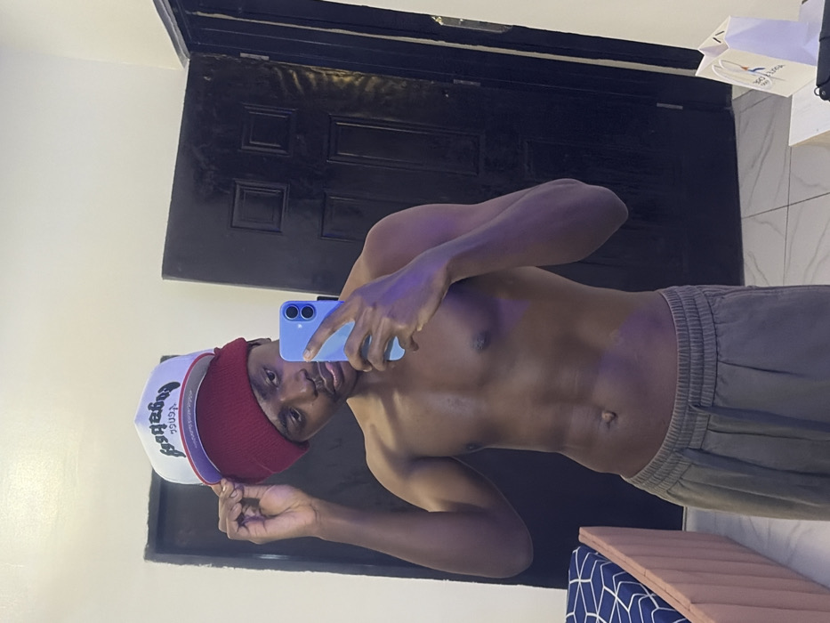 A shirtless man wearing gray sweatpants and a maroon and white baseball cap takes a selfie using a light blue phone. He poses against a background that features a black door and tiled walls. The man's physique is visible, including his chest, stomach, and arms. A white shopping bag is partially visible near the top corner of the shot. The image is rotated, displaying the subject sideways.