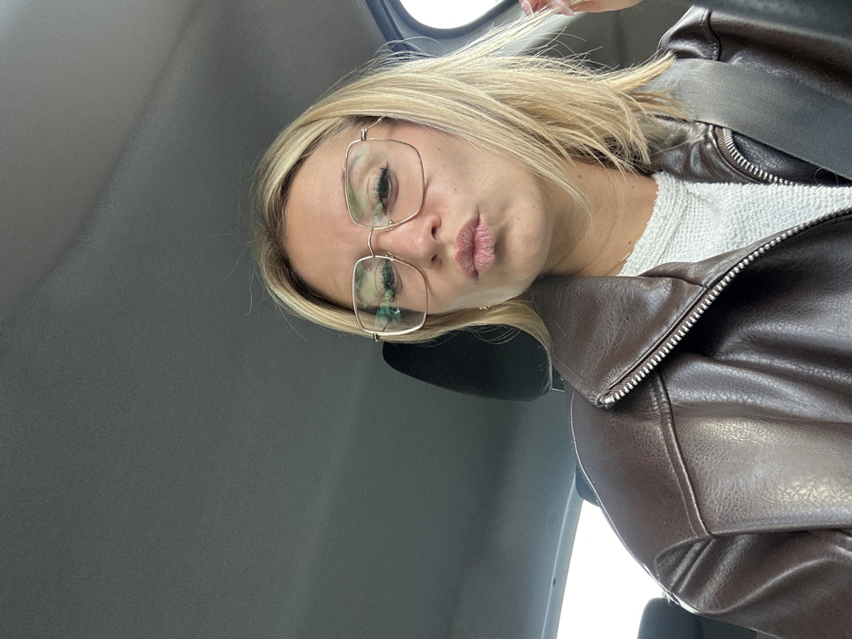 A female individual is depicted from the mid-chest up, seated inside a vehicle in Villeneuve-Loubet, France. She has blonde, shoulder-length hair and wears large, square-framed glasses with light-colored metal frames. Her lips are pursed in a puckering expression. She is clad in a dark brown, zippered jacket, possibly of faux leather, over a light-colored, textured top, and a black seatbelt crosses her upper torso. The vehicle's dark grey headliner and a black headrest are visible in the background. A bright, undifferentiated light is visible through a window on the right side of the frame.