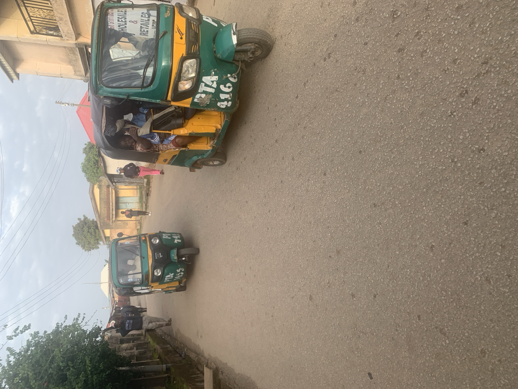 An outdoor, eye-level, medium shot captures a street scene in Jos, Nigeria, with a focus on two auto-rickshaws, also known as "Keke Napep." The auto-rickshaws are a common mode of transport in Nigeria. The rickshaws are green and yellow, and both have visible license plates with PTA markings. The auto-rickshaw in the foreground has the license plate PTA 4166. It is carrying several passengers, including a woman in the passenger seat who is giving a thumbs-up. The rickshaw in the background has the license plate PTA 1455. A man in a grey hooded sweatshirt and khaki pants is walking away from the camera and gesturing with his right hand raised. Behind him, three other men in dark suits are walking down the street. The street is paved with asphalt and is lined with buildings. The sky is overcast.