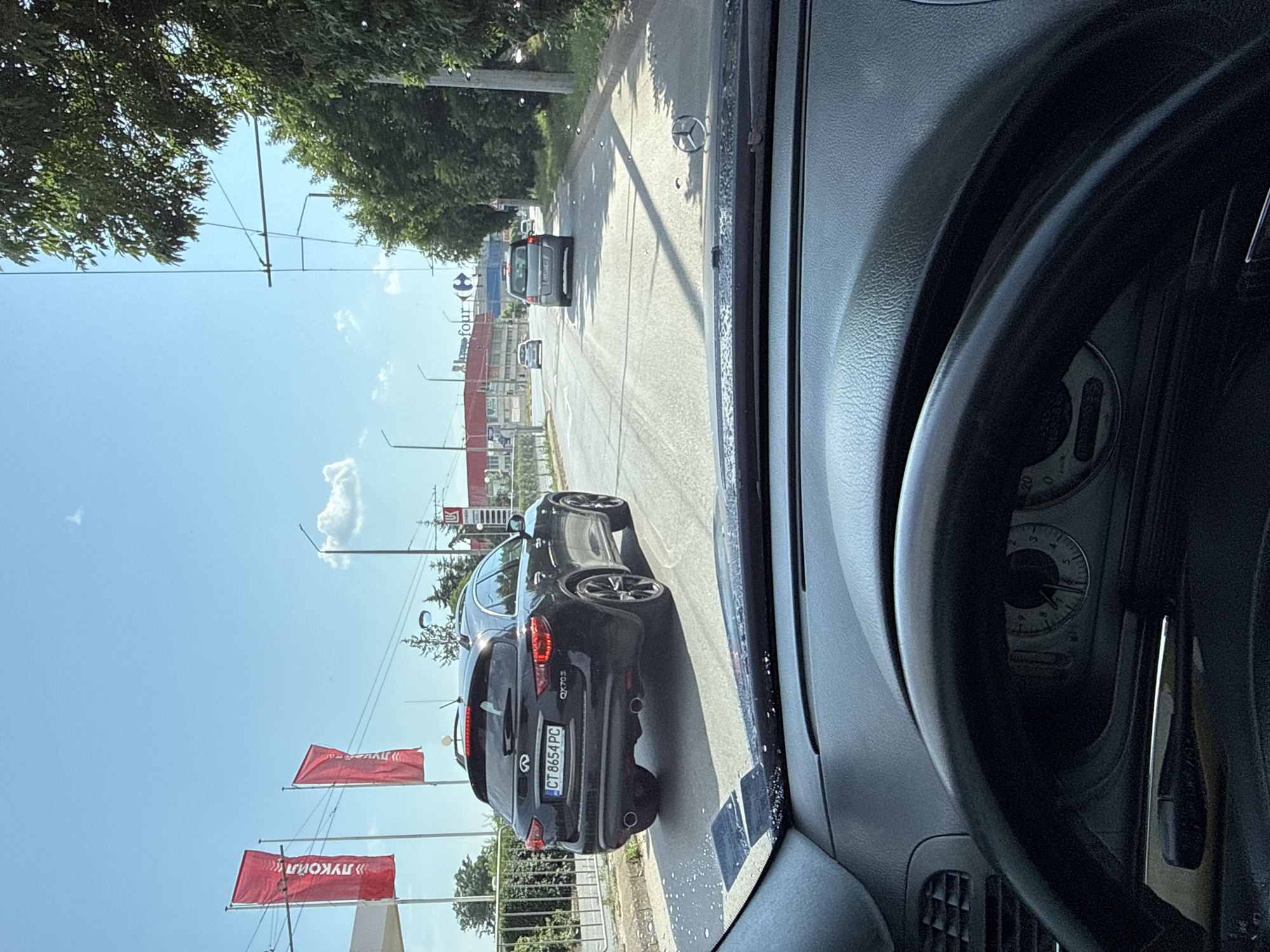 The image depicts an urban street scene in Stara Zagora, Bulgaria, captured from the driver's perspective inside a Mercedes-Benz vehicle.
In the immediate foreground, the top of a dark gray dashboard and a section of a black steering wheel are visible. A Mercedes-Benz star emblem is present on the dashboard, and several water droplets are scattered across the windshield.
The midground features an asphalt road with visible white lane markings. A black Infiniti QX70S SUV, bearing Bulgarian license plate "CT 8654 PC" (CT indicating Stara Zagora), is directly ahead, moving away from the viewer. Its rear brake lights are illuminated. To the left of the road, a metal fence separates the roadway from a commercial area. To the right, a concrete curb borders a strip of dense green foliage. Further down the road, a grey minivan/SUV and a white sedan are also in motion.
The background shows a blue sky with a few scattered white clouds. On the left, two red flags with "ЛУКОЙЛ" (Lukoil) in white Cyrillic script are displayed, indicating a Lukoil gas station. Prominently in the center, a large red building with "four" and the distinctive blue and red Carrefour logo is visible, signifying a Carrefour supermarket. Various utility poles, streetlights, and overhead wires are present along the road, interspersed with green trees.
The scene is illuminated by bright daylight, suggesting a sunny day. No individuals are explicitly visible, but the presence of moving vehicles implies human activity.