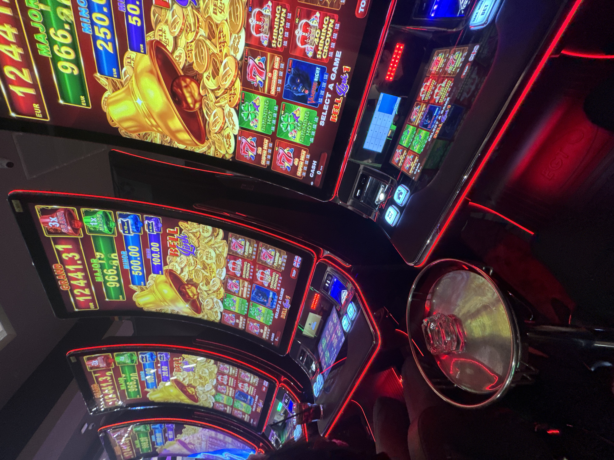 The image shows a row of modern slot machines, likely in a casino. The slot machines feature bright, colorful displays with themes involving gold coins and classic casino symbols. Some of the visible games include "Burning Hot", "Shining Crown", and "Vampire Night." Jackpot amounts for "Grand," "Major," "Minor," and "Bell Link" are displayed on the screens, along with the currency "EUR."
In the foreground, the edge of a chair is visible, suggesting someone is sitting in front of one of the machines. Red LED lighting accents the machines, adding to the ambiance of the environment. The image was likely taken in Plovdiv, Bulgaria.