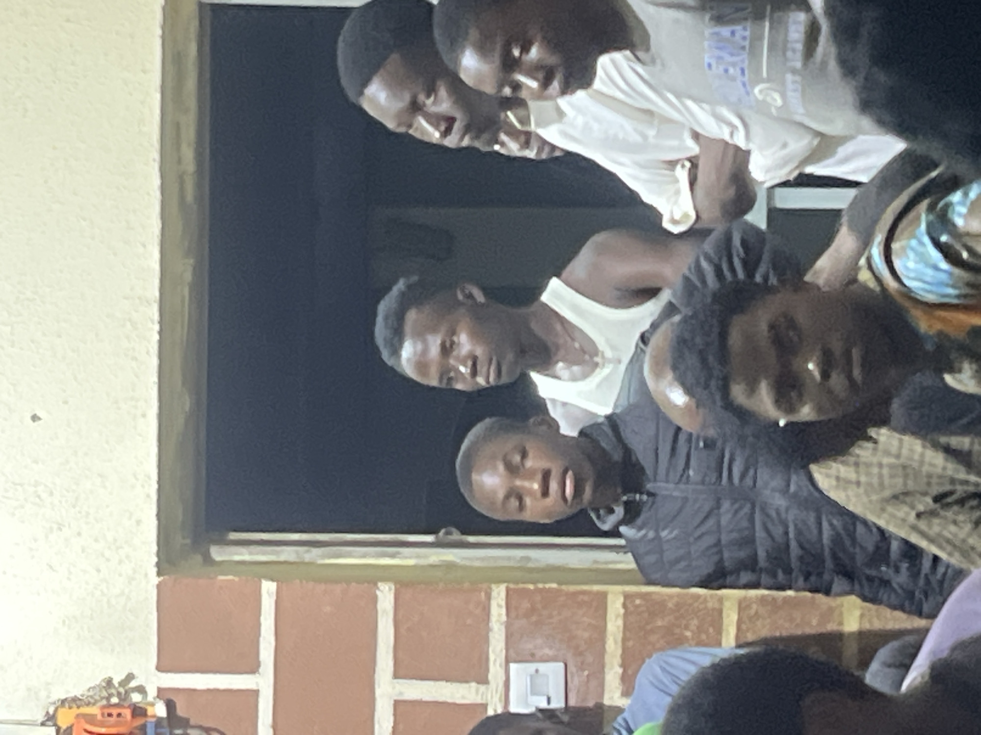 A group of young Black men are gathered in what appears to be the doorway of a building in Jos, Nigeria. The lighting suggests it is evening or indoors. The men are of varying ages, appearing to be teenagers and young adults.
On the far left, a person is partially visible, wearing a light blue shirt. To their right, a young man in a black puffer jacket looks directly at the camera. Behind him, another young man wears a white tank top and a cross necklace, looking slightly to the left. Further to the right, a man in a light-colored long-sleeved shirt with rolled-up sleeves is also looking to the left, his arms crossed. In the foreground, to the right, another young man wears a gray t-shirt with blue lettering that reads "KIDEMAN" and a phrase below it. Other individuals are partially visible in the foreground and background, their faces and clothing obscured.
The environment shows a brick wall on the left with white horizontal trim and a light switch. The doorway leads into a dark interior. The overall impression is one of a casual gathering or meeting.