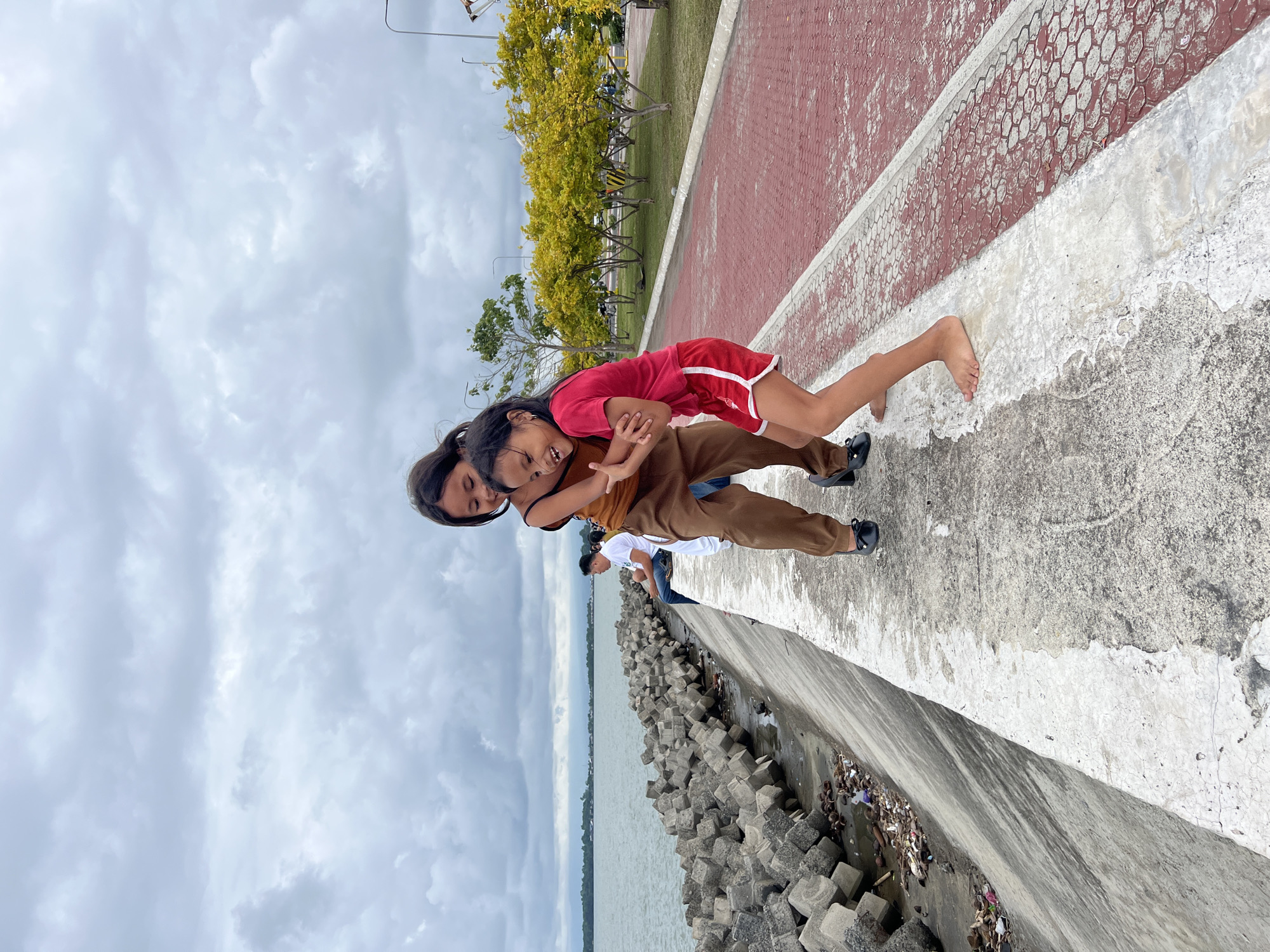 Two young girls are playing on a concrete promenade next to a body of water. The girl on the left is wearing a brown top and brown pants, and is embracing the girl on the right. The girl on the right is wearing a red t-shirt and red shorts, and is laughing. The promenade is paved with red hexagonal tiles and bordered by a white line. To the left of the promenade is a concrete embankment with large concrete blocks and some debris. In the background, there is a body of water, a distant shoreline, and a row of trees with yellow leaves. The sky is overcast with gray clouds. In the middle ground, to the left, a man is sitting on the embankment, facing away from the camera, wearing a white t-shirt and blue jeans. This scene is located at the Davao City coastal promenade, a popular recreational area in Davao City, Philippines.