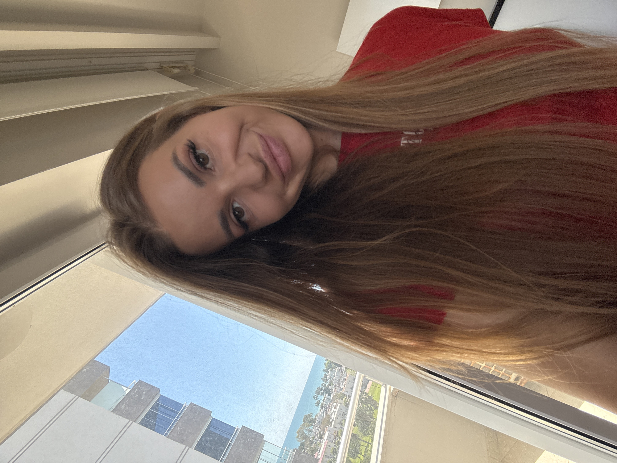 A woman with long, straight, light brown hair is facing the camera with a slight smile. She is wearing a red top. Behind her, a window reveals a view of a city, likely Erdemli, Turkey, based on the coastal setting. Visible are buildings, trees, the Mediterranean Sea, and a clear blue sky. The window frame is white, and there are window blinds visible at the top of the frame. The perspective is slightly tilted.