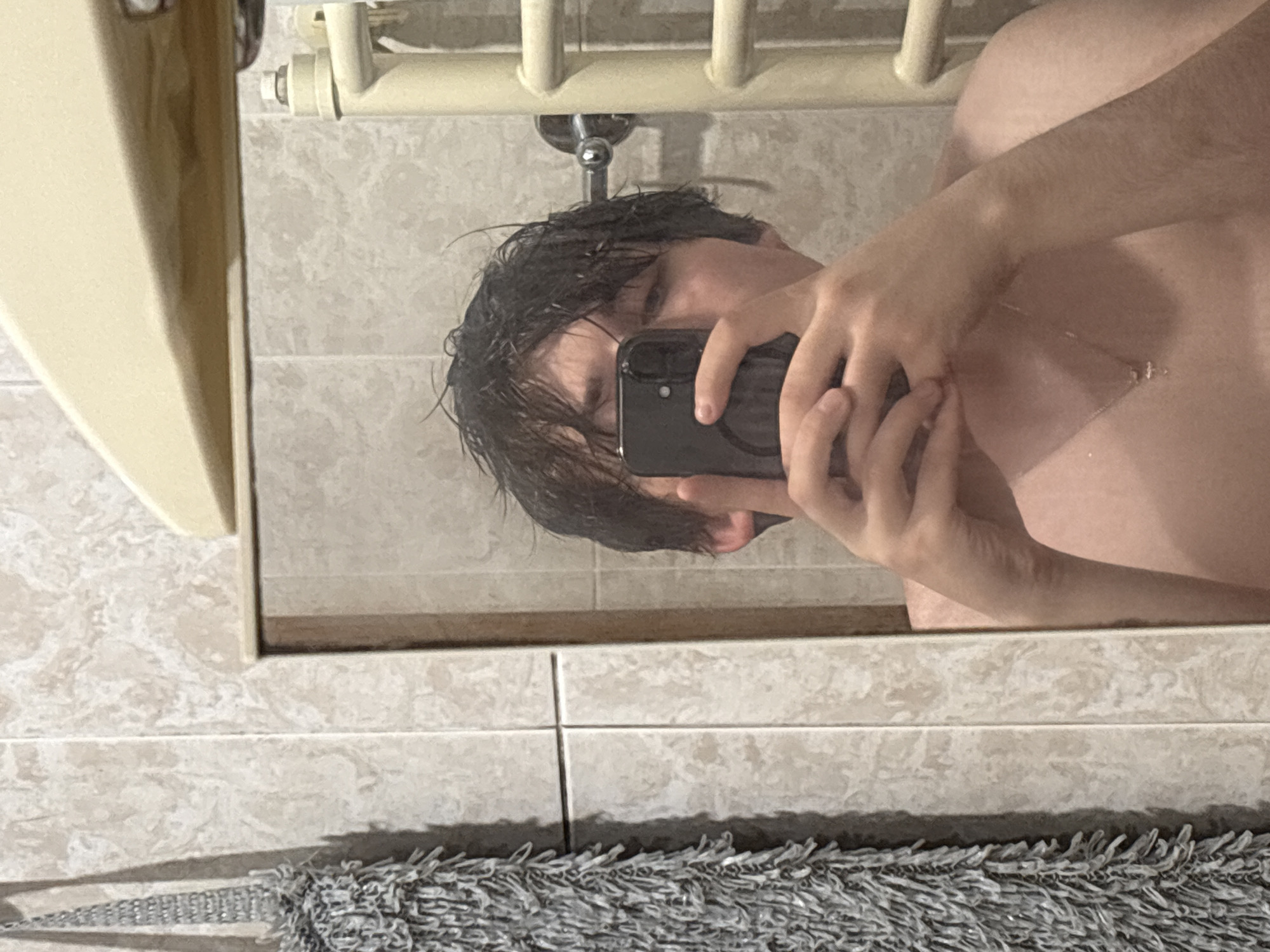 A young person with dark, wet hair is taking a mirror selfie in a bathroom. They are holding a black smartphone in front of their face, obscuring most of their features. The person is wearing a thin gold necklace with a small cross pendant. The background consists of beige tiled walls, a white towel hanging on the left, and a beige towel rack on the right. The scene appears to be in a residential bathroom. There is no information to determine the specific location in Dnipro, Ukraine.