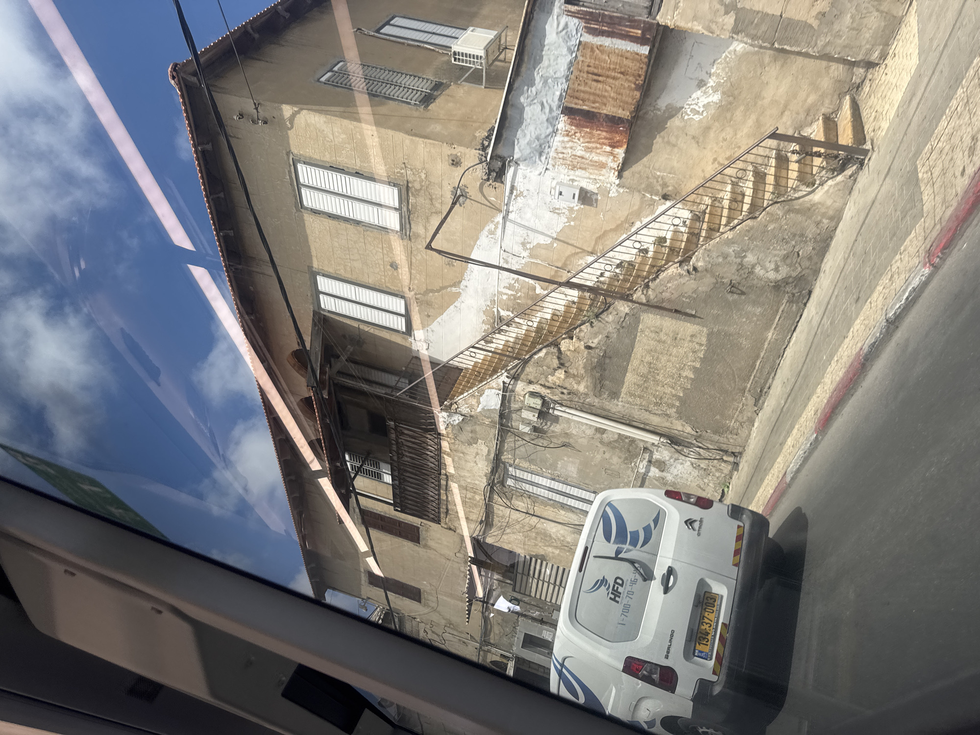 A medium-angle shot depicts an urban scene in Tel Aviv, Israel, viewed through a car window. The focus is on a weathered, multi-story building with a visible external staircase. The building's facade is characterized by a mix of light-colored paint and exposed concrete, showing signs of age and disrepair. Several windows, some with metal shutters, are visible. A white Citroen Berlingo van, bearing the branding "HFD" and a phone number, is parked on the street adjacent to the building. The vehicle's license plate is partly obscured but appears to be Israeli. The sky is visible in the window's reflection, suggesting a sunny day. Power lines are strung across the building.