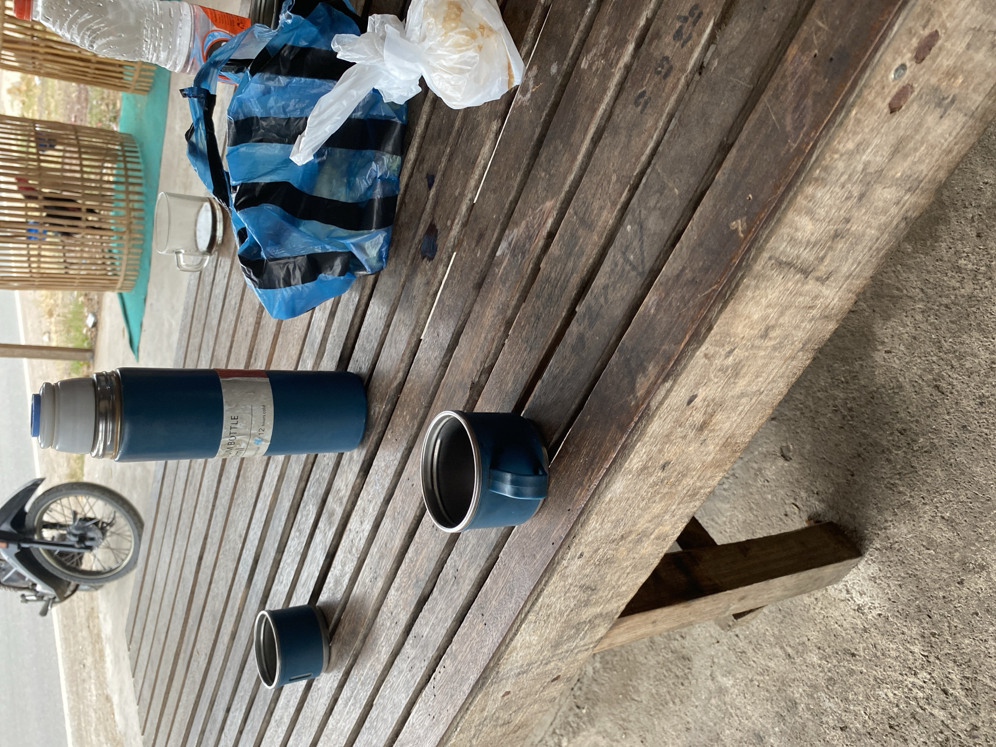 This is a medium-angle, eye-level shot of a weathered wooden table with several items on it. The table is outdoors, as indicated by the rough concrete ground and the presence of a motorcycle in the background. In the foreground, there are two blue metal cups with handles, one is upside down. To the right of the cups is a tall, blue vacuum flask. Further to the right, there is a clear glass mug with a dark liquid, likely coffee, and behind it, a clear plastic bottle containing a pink liquid, possibly a beverage. A crumpled blue and black striped plastic bag is also visible, as is a white plastic bag containing some light-colored food items. In the background, a motorcycle with a partially deflated tire is parked on a paved surface. Beyond the motorcycle, there are two bamboo structures, one of which appears to contain a rooster. The lighting suggests it is daytime, possibly late morning or early afternoon. The scene is set in an outdoor eating or resting area in Bayu, Indonesia.