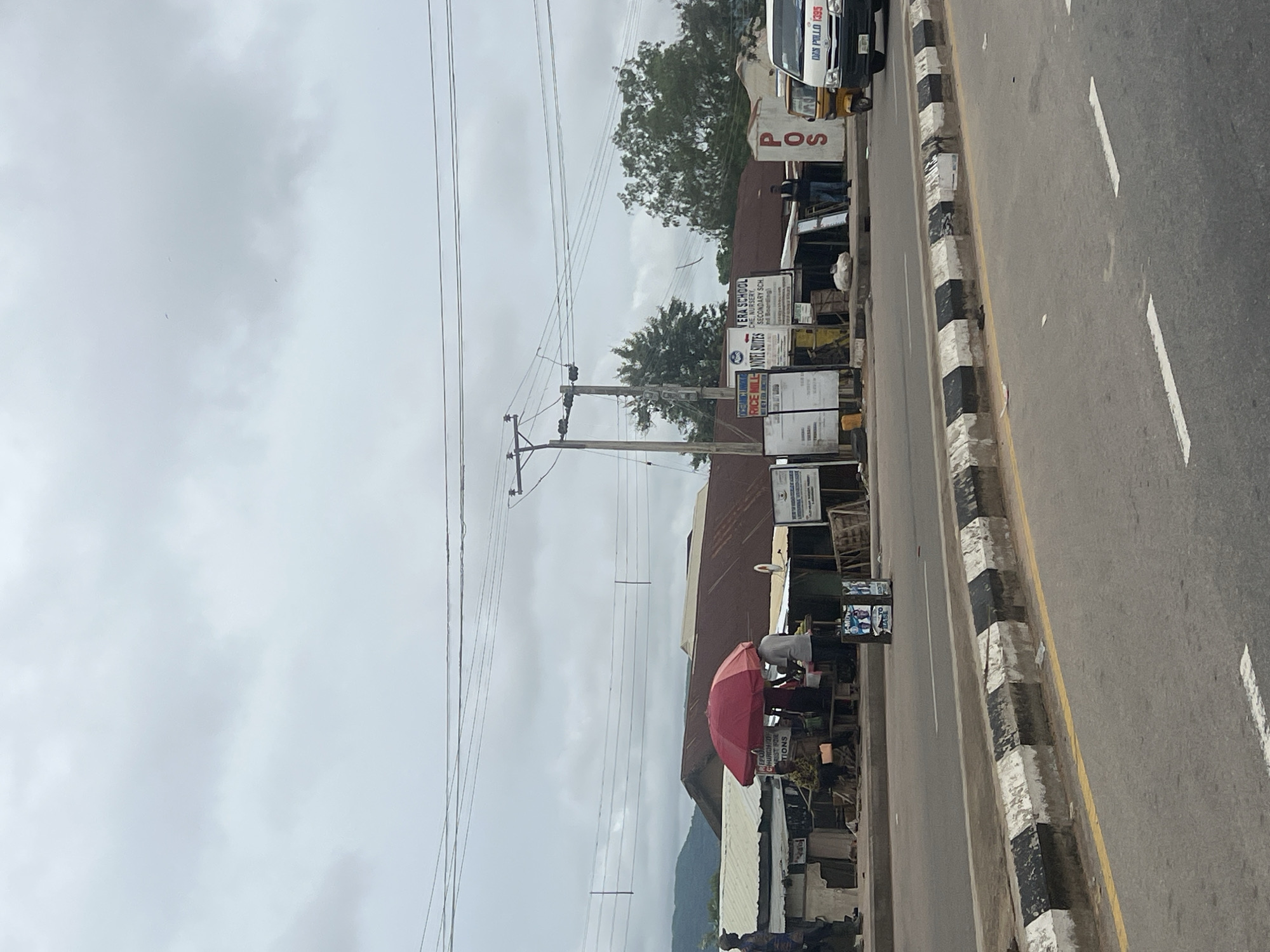 The image captures a roadside view in Jalingo, Nigeria, under an overcast sky. A street market with several stalls is visible along the side of a road. One stall features a prominent red umbrella shielding a vendor and a customer. Various goods, including produce, are displayed. In the background, buildings with corrugated metal roofs are present. Signs indicating businesses such as "RICE MILL," "NOVEL SUITES," and "VERA SCHOOL" are displayed. A "POS" sign is mounted on a pole. Several vehicles, including a white van with "DAN POLLO 1395" on its side and a yellow motorized tricycle, are parked or driving on the road. A few individuals are seen in and around the market stalls. The road has distinct black and white painted concrete barriers along its edge, with a yellow line marking the lane. Overhead power lines crisscross the sky. The scene suggests a busy commercial area in the city.