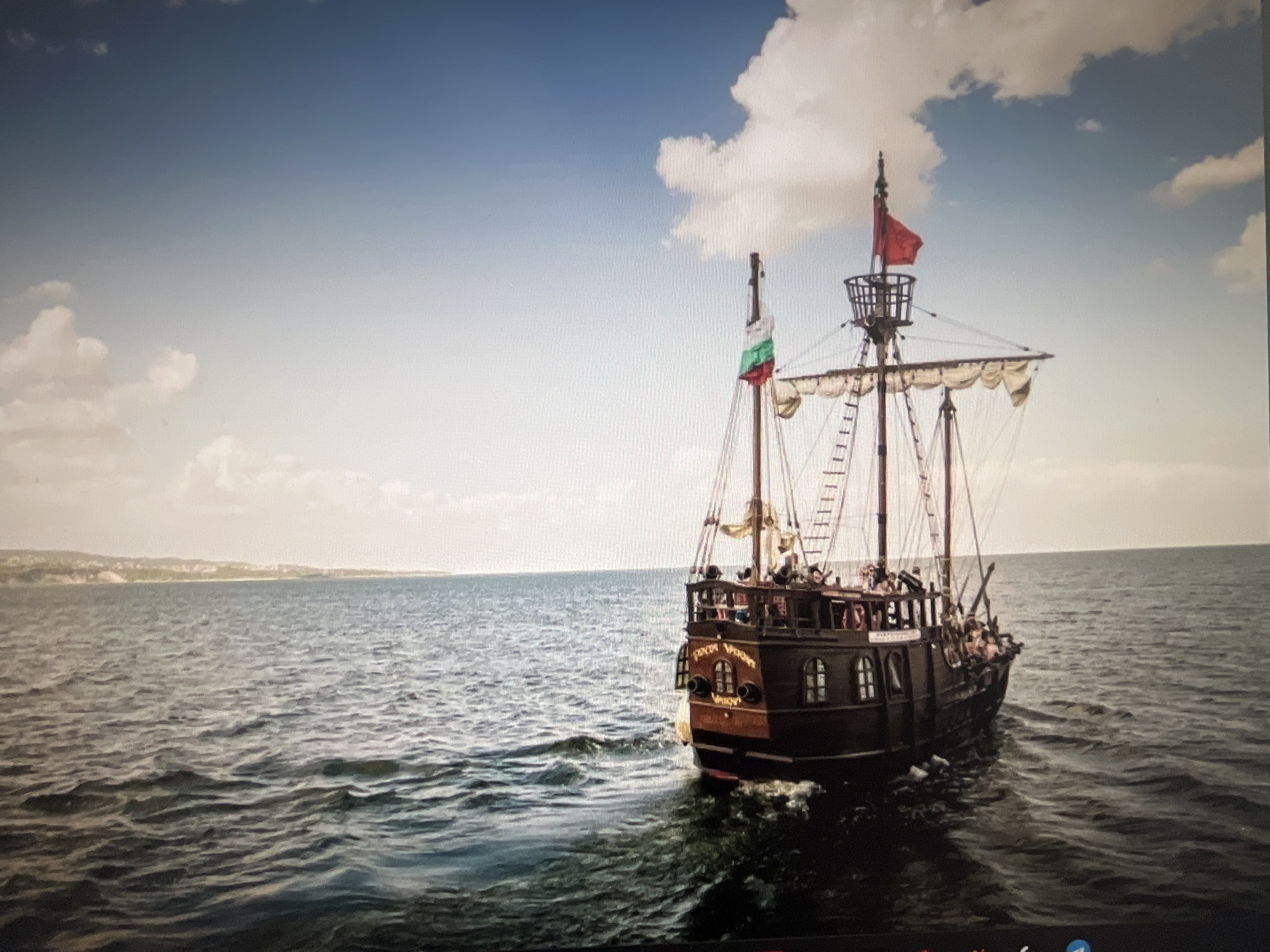 A wooden pirate ship sails on the Black Sea, with a coastline visible in the distance. The ship has two masts with sails, and a red flag flies from the top of the main mast. A Bulgarian flag is also visible on one of the masts. There are people on deck, and the ship is decorated with carved wooden panels. The sea is choppy, and the sky is partly cloudy. The ship appears to be the "Sea Dreamer" pirate ship, which is a tourist attraction in Varna, Bulgaria.