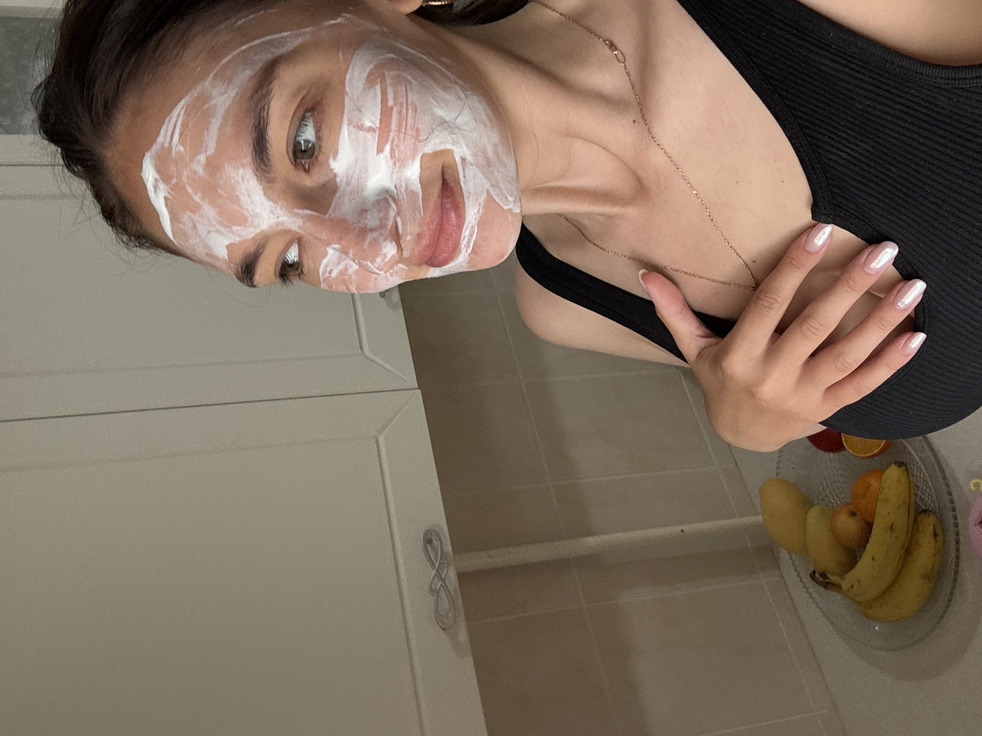 A woman with a white facial mask poses for a selfie in an indoor setting. She is wearing a black tank top and a delicate gold necklace. Her manicured hand rests near her chest. A fruit bowl containing bananas, mango, oranges, and other fruit sits on a tiled surface. The image also includes a portion of a white cabinet with a silver handle. There is no indication that this image was taken in Sochi, Russia.