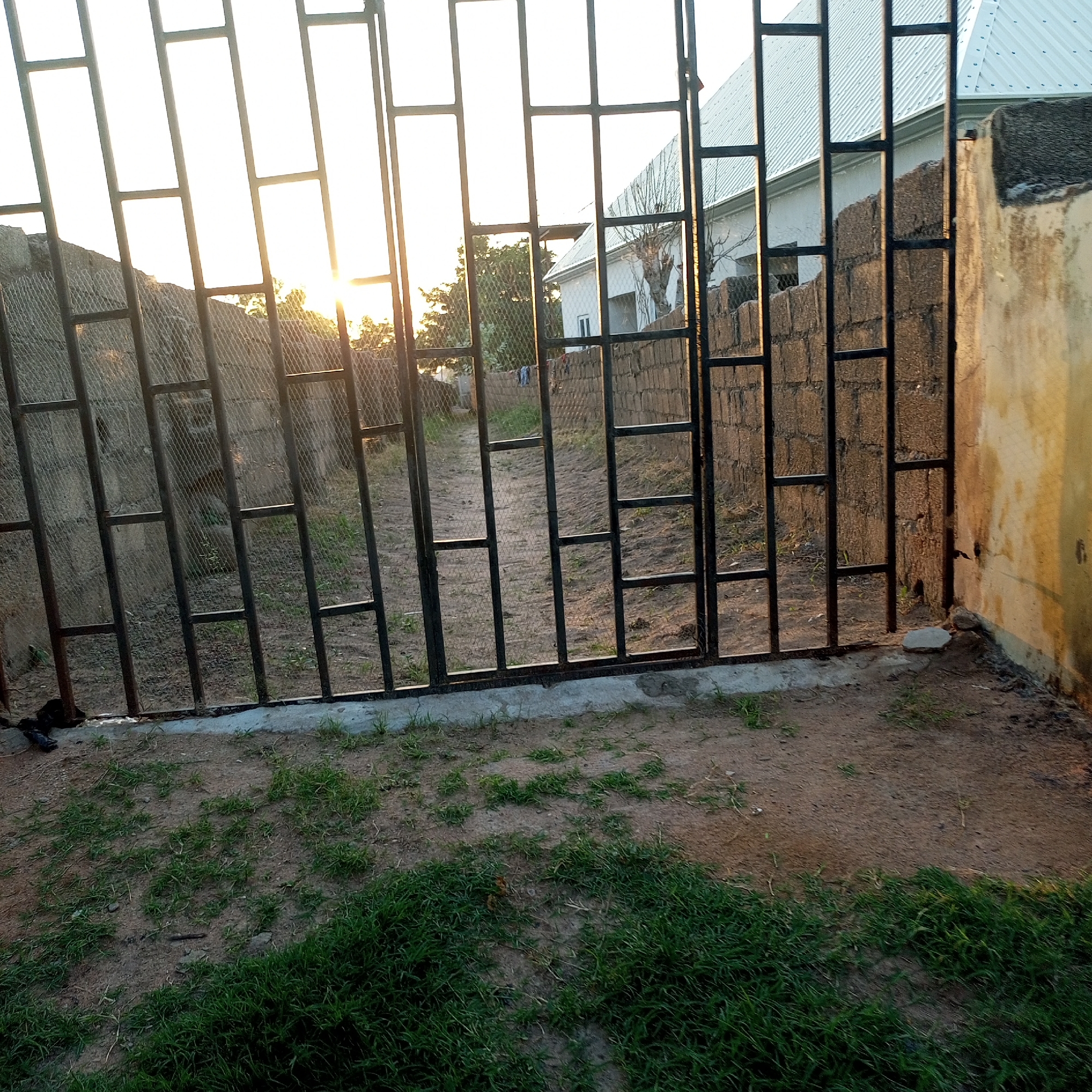 The image depicts a street view through a metal gate, with the sun setting in the background, casting a warm glow on the scene. The gate, made of dark metal bars, forms a grid pattern across the foreground. Beyond the gate, a dirt path leads into a sparsely vegetated area, where a building with a corrugated metal roof is visible. The walls of the building and adjacent structures are constructed from cinder blocks and plastered surfaces, some of which are weathered and discolored. The ground is a mix of dirt and patches of dry grass. In the distance, trees can be seen. The overall impression is that of a residential area in Jalingo, Nigeria, during a late afternoon or early evening.
