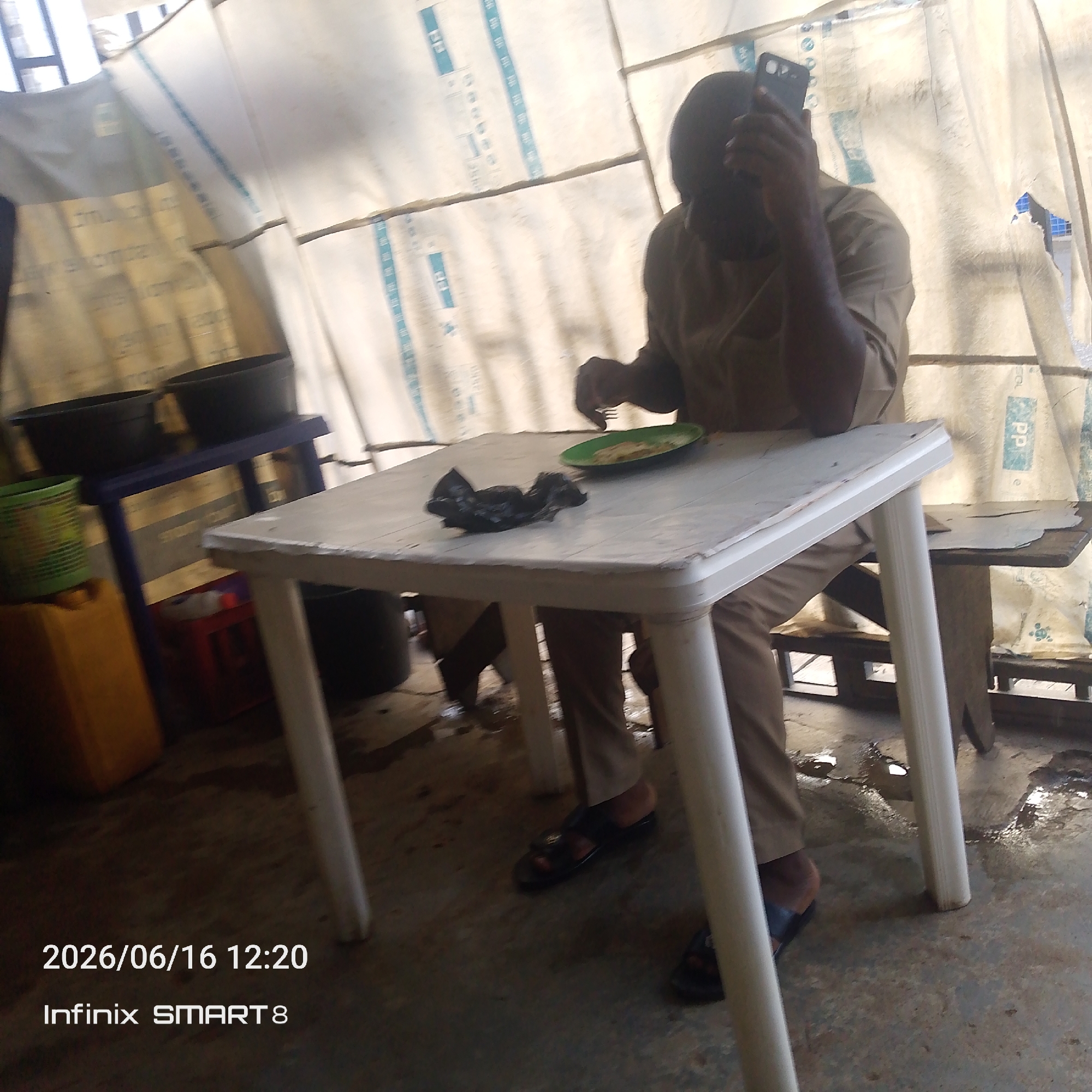 In this image, a person is sitting at a white plastic table, holding a black smartphone. The person is wearing a light brown collared shirt and matching pants, and black sandals. On the table, there is a green plate with food, and a crumpled black object that appears to be a plastic bag.
The scene is set outdoors, under a makeshift shelter made of white plastic sheeting. The sheeting has blue-striped patterns printed on it. The ground is concrete and appears wet in places. To the left of the table, there is a blue shelf with two black pots on it, and a green basket and a yellow container. A small red toy is visible beneath the shelf. In the background, more of the plastic sheeting and some wooden structures are visible.
The image captures a moment in time, likely during the day, given the lighting. The overall impression is of a simple, perhaps improvised, outdoor eating or resting area. The person appears focused on their phone.
Visible text on the image includes "2026/06/16 12:20" and "Infinix SMART 8" in the bottom left corner.