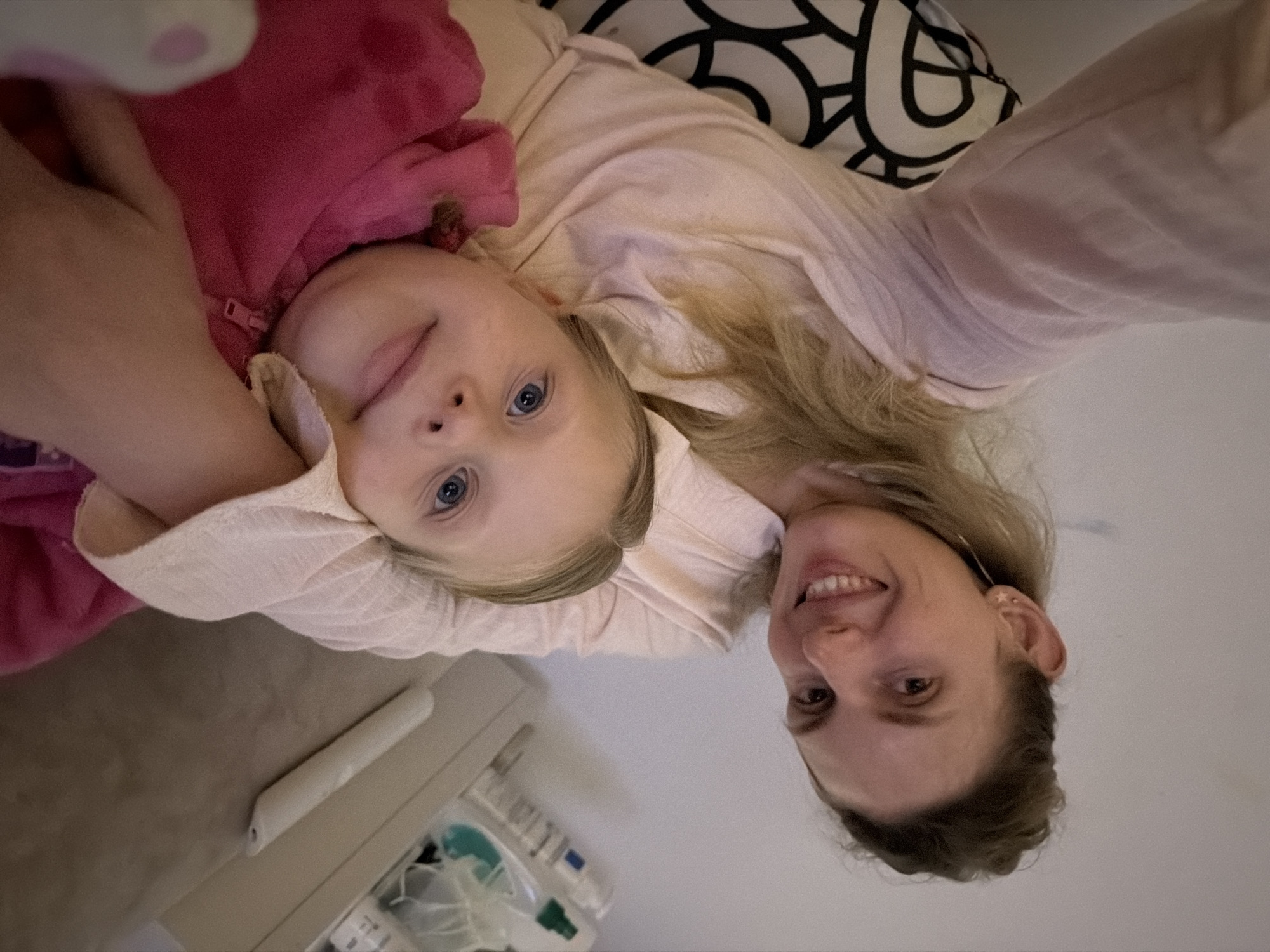 A woman and a young child are lying down together, possibly on a bed or soft surface. The child is wearing a pink jacket, and the woman is in a light-colored shirt. The background includes a pillow with a black and white pattern. There is also a white object, possibly medical equipment, in the lower left corner of the frame. The location in the city of Budva, Montenegro cannot be determined from this image.