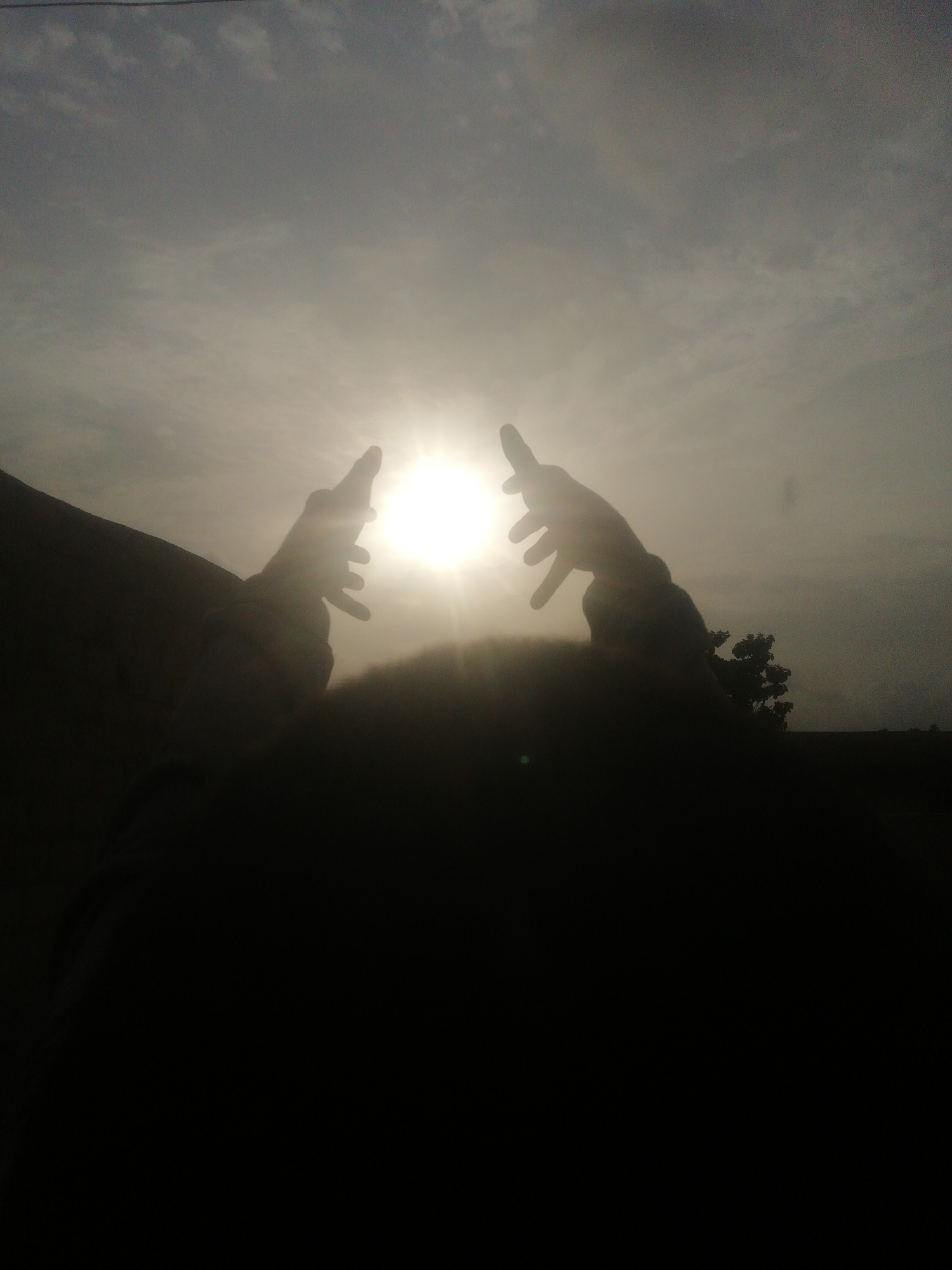 The image shows two hands reaching up towards the sun in an area with a cloudy sky. The foreground is dark, obscuring details, but it appears to be a person. The environment includes some vegetation and structures. The city and specific location are not discernible.