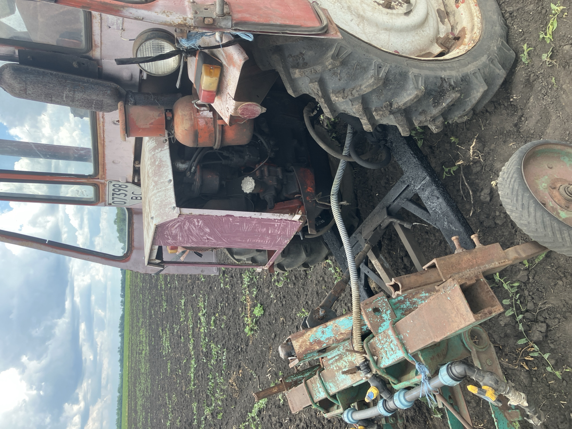 The image shows a red tractor with a planting attachment in a field. The tractor appears weathered and shows signs of rust. It has a license plate that reads "07398 BC". The planting attachment is green and rusty, with blue pipes and yellow valves. The field is plowed and appears to be recently worked. The sky is cloudy. The location is a field in the city of Berdianka, Ukraine.