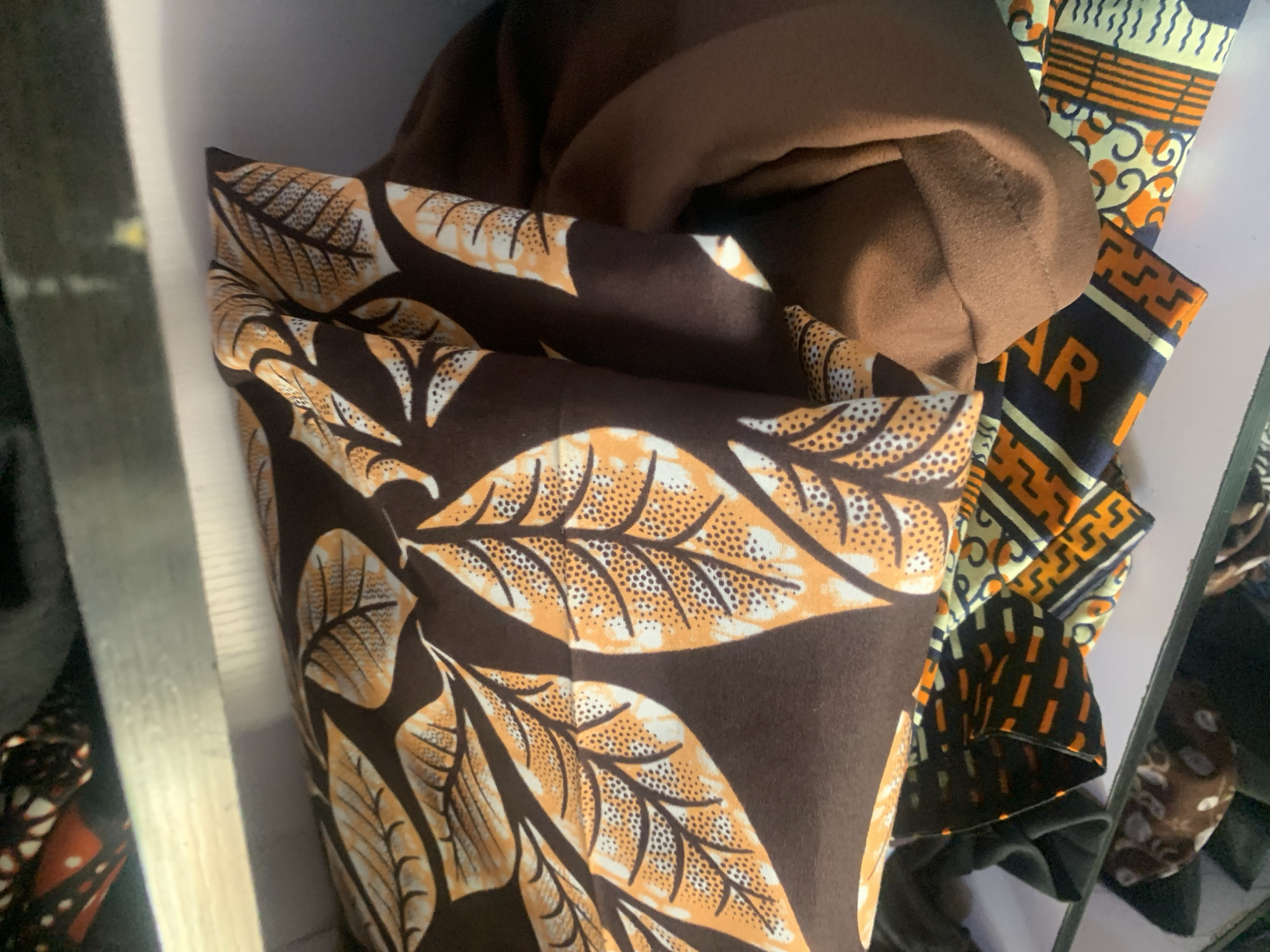 In Jauro Yinu, Nigeria, a close-up shot focuses on a stack of folded fabrics. The top fabric displays a bold pattern of large, stylized leaves in orange and white against a dark brown background. The leaves are detailed with a textured fill and prominent veins. Beneath this, another fabric is visible, exhibiting a geometric pattern with black, orange, and yellow elements. The textures suggest these are textiles, possibly intended for clothing or home decor. The lighting is diffused, highlighting the rich colors and patterns of the fabrics. The environment appears to be indoors, possibly a shop or storage area, with a shelf visible above the stacked items.