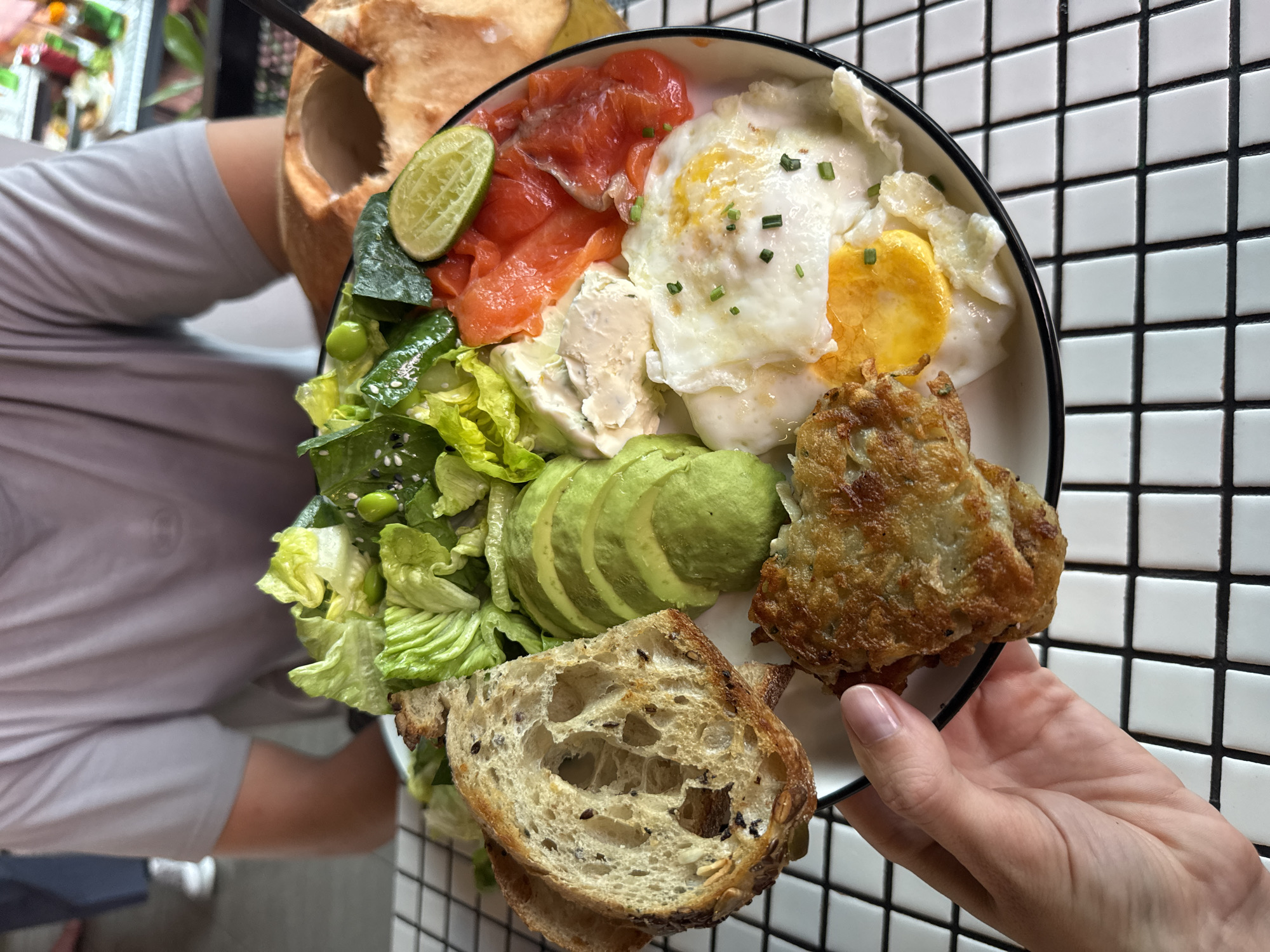 A person is holding a plate of food containing slices of salmon, an egg, avocado, a piece of bread, a piece of fried food, and salad. A coconut drink is behind the plate. The plate is being held in front of a tiled wall, which is likely indoors at a restaurant in North Kuta, Indonesia.