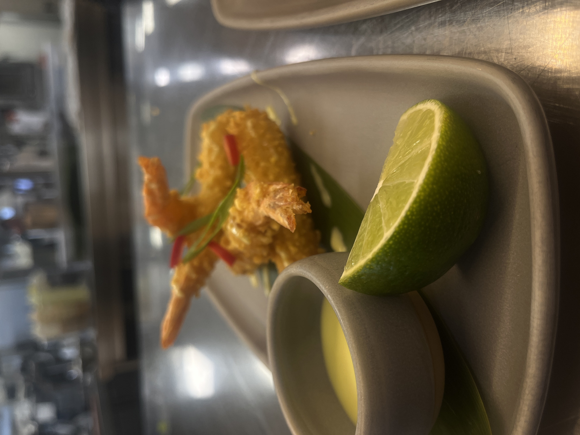 The image displays a close-up, high-angle shot of a plated food item, likely an appetizer, presented on a light gray, irregularly shaped dish. The main subjects are several pieces of golden-brown, breaded shrimp, accompanied by a bright green lime wedge and a small bowl of pale yellow dipping sauce.
The shrimp appear to be fried and are garnished with slender red chili strips and green herbs, possibly spring onions or chives. The lime wedge is cut, revealing its juicy interior, and is positioned beside the small, matching gray bowl containing the creamy sauce. Another, larger plate is partially visible behind the main dish, along with hints of green leaves as part of the garnish.
The background is out of focus, suggesting a shallow depth of field. It shows blurred reflections on a metallic surface, characteristic of a commercial kitchen or restaurant environment, possibly a stainless steel countertop or appliance. The lighting is artificial, illuminating the food brightly. There are no people visible, nor any discernible text. The image does not contain specific visual cues to indicate its location in Sofia, Bulgaria.