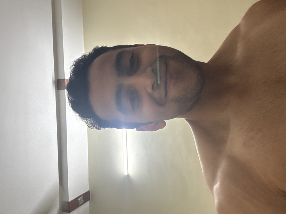 A man's face and bare chest occupy most of the frame. His dark hair is visible at the top left. The man is smiling slightly, with light reflecting off his face, possibly from a light fixture behind the camera. A white wall is on the left side of the image, with dark brown trim and a light beige wall in the background. The image is taken from a low angle, looking up at the man.