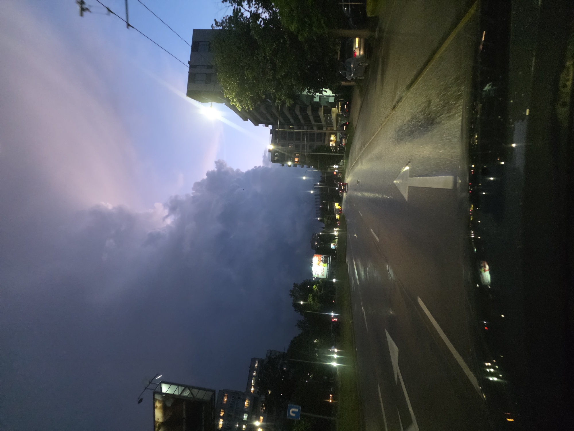 The image depicts a street scene in Varna, Bulgaria, at dusk or night, likely after rain, as indicated by the wet pavement. The view is oriented along a road, with buildings visible on both sides, illuminated by streetlights and lights from within the structures. A large, dark cloud formation dominates the sky on the left side of the image, contrasting with the remaining daylight. The road itself features painted directional arrows and lane markings, with reflections of the lights creating a shimmering effect. There are also billboards and other signs visible, adding to the urban landscape. There are cars visible along the road.