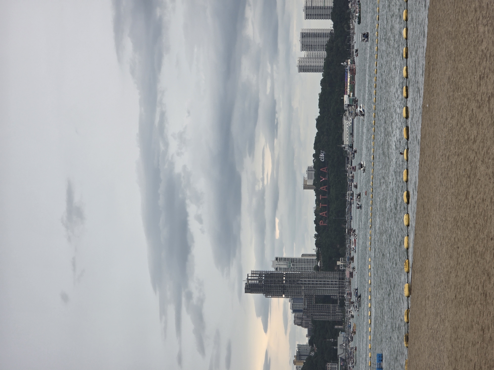 This image depicts a wide shot of Pattaya City, Thailand, from a beach. The foreground features a sandy beach with yellow buoys marking a separation in the water. The sea is visible in the midground, with numerous boats of various sizes scattered across its surface. In the background, the Pattaya City skyline is prominent, characterized by a cluster of tall buildings, including a high-rise under construction. A large sign on a hill reads "PATTAYA city." The sky is overcast with a mix of gray and white clouds.