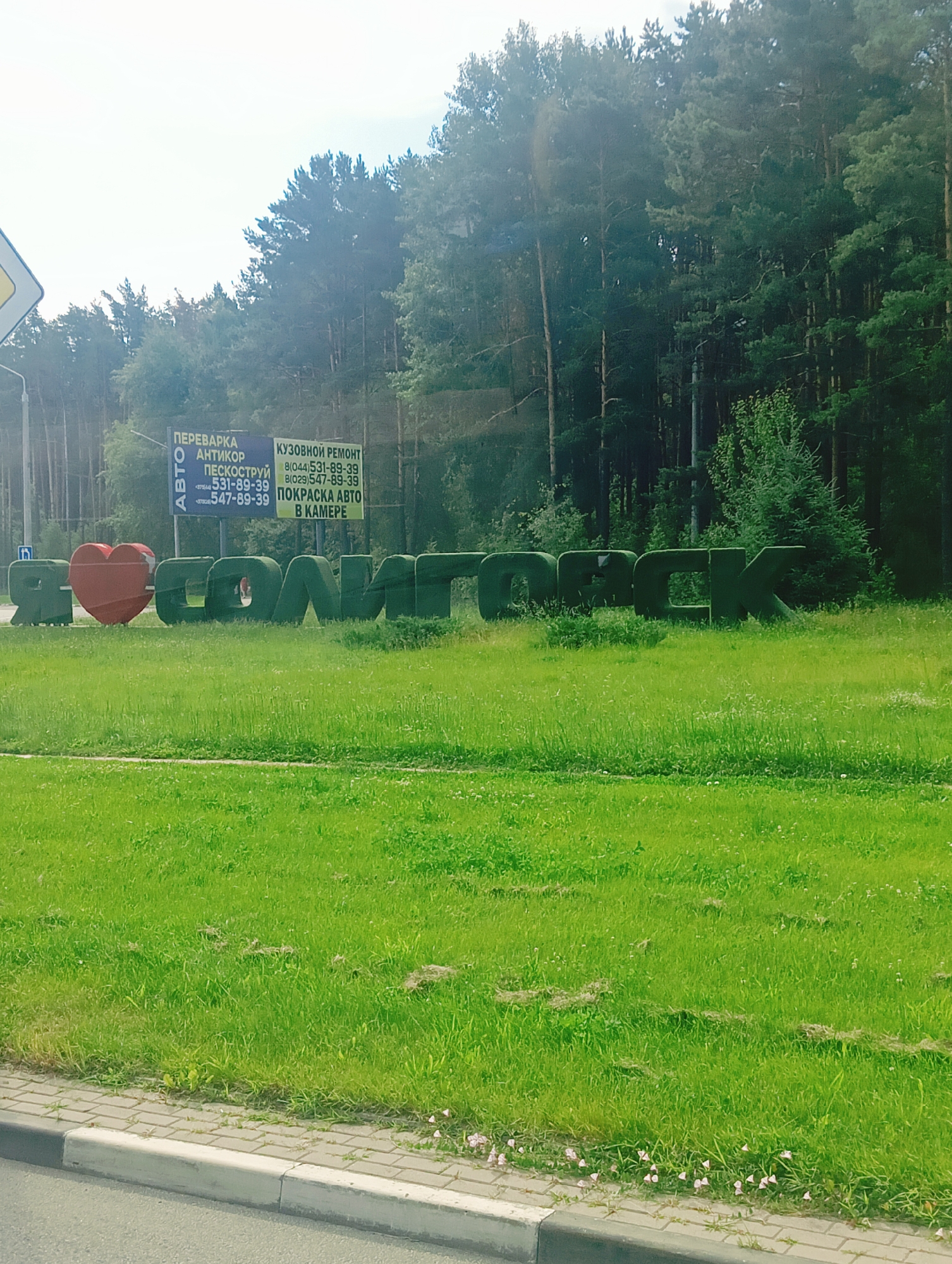 The image depicts a lush green lawn with a large, red heart sculpture and green, topiary-style letters spelling out "Я ЛЮБЛЮ СОЛИГОРСК" (I Love Salihorsk) in the foreground. Behind the sculptures, a dense forest of tall pine trees forms a natural backdrop. A bright, overcast sky fills the upper portion of the image. To the left, a yellow traffic sign is partially visible, and a utility pole stands within the forest. Two billboards are mounted on poles behind the sculptures; one advertises car services like "ПЕРЕВАРКА АНТИКОР" (Welding Anti-corrosion) and "ПЕСКОСТРУЙ" (Sandblasting) with phone numbers, while the other promotes "КУЗОВНОЙ РЕМОНТ" (Body Repair), "ПОКРАСКА АВТО В КАМЕРЕ" (Car Painting in a Chamber), also with phone numbers. The foreground also shows a paved sidewalk and a section of asphalt road. The overall scene captures a welcoming message to the city of Salihorsk, Belarus, set against a natural, wooded environment.