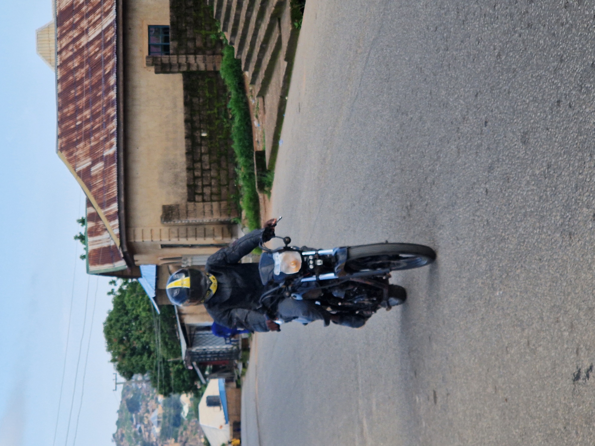 A person wearing a black motorcycle jacket and helmet with yellow accents is riding a black motorcycle on a paved road. The motorcycle has a visible headlight and front wheel. In the background, there are buildings with corrugated metal roofs, some with light-colored walls and brick accents. A stone wall with greenery behind it and a set of concrete stairs are also visible. The environment appears to be an urban or semi-urban setting in Kabong, Nigeria, with hints of hilly terrain in the distance. The sky is overcast.