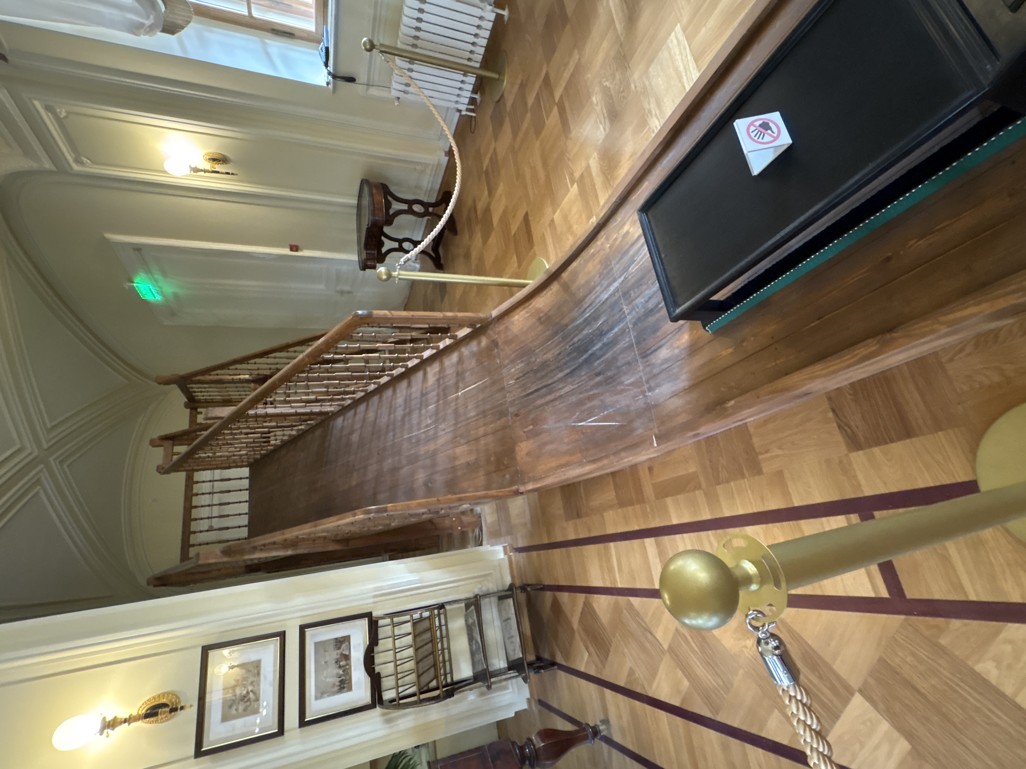 In the interior of the Gatchina Palace, a wooden slide descends from a landing partway up a staircase. The slide is made of dark wood and appears well-worn. A black mat with a prohibition sign is at the bottom of the slide. The floor is made of parquet flooring with contrasting bands. A brass stanchion with a rope barrier is in the foreground. The room has a high ceiling with decorative molding. A vintage ladder-like shelf with books or papers is to the left of the slide. Two framed pictures hang on the wall above the shelf. A small, ornate table stands near the top of the slide. A radiator is visible on the right wall. An illuminated green "EXIT" sign is mounted on a white door in the background.