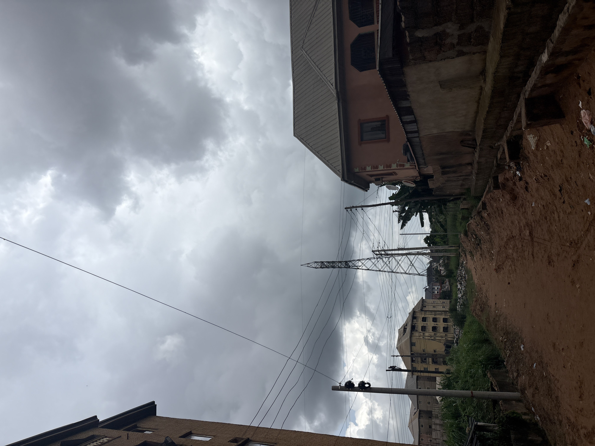 The media file depicts an outdoor scene in Nkpor, Nigeria, under a heavily overcast sky. The view is primarily upward, showcasing a dramatic sky filled with dark, moody clouds, suggesting imminent rain or recent downpour.
Prominently featured are a series of power lines and a large, lattice-style electricity pylon, stretching across the frame and disappearing into the dense cloud cover. These are juxtaposed with buildings, two of which are partially visible. On the right side of the frame, a rough, reddish-brown dirt embankment is visible, with some scattered debris suggesting a less-maintained area. Above this embankment, a building with a pinkish-tan facade and a metal roof is partially seen. Further into the scene, a multi-story building with numerous windows, some boarded up or dark, stands out. There is also another building visible in the background, appearing somewhat weathered.
The overall atmosphere is somber and slightly dramatic due to the intense cloud formation. The weather appears to be wet or about to become wet, contributing to a sense of gloom. No people are visible in the frame. The presence of the power lines and pylon, along with the style of the buildings, are common elements in urban or semi-urban environments in Nigeria. The time of day is difficult to ascertain precisely due to the overcast conditions, but the dim light suggests it could be late afternoon or a particularly dark, cloudy morning.