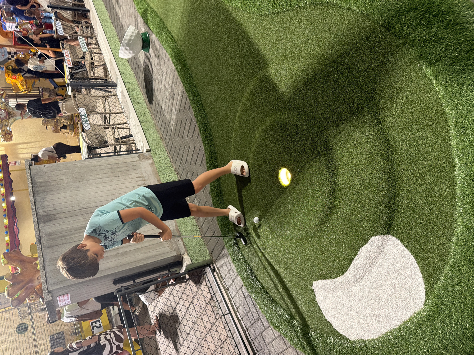 In Gatteo, Italy, at a mini-golf course, a young boy with brown hair is focused on hitting a white golf ball with a black putter. He is wearing a light blue t-shirt with a tropical print and black shorts, and white slide sandals. The boy stands on the green artificial turf, with a sand trap to his left and a hole with a glowing light to his right. In the background, other people are visible near a fence and a row of tables and chairs. The setting appears to be an amusement area with carnival-like attractions in the distance.