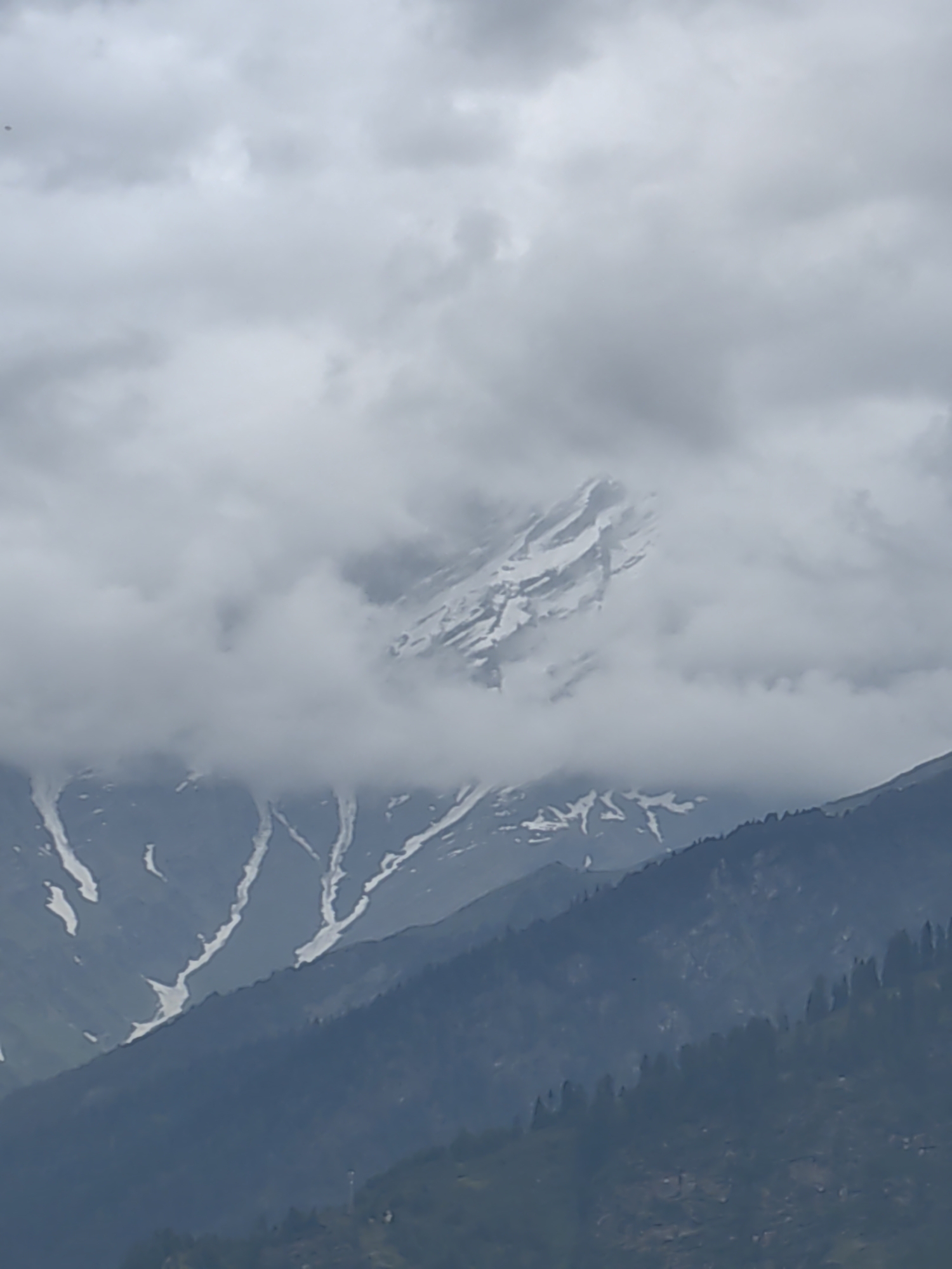 A photograph taken from Vashisht, India, captures a cloudy, overcast day with a dramatic mountain landscape. The foreground features dark, densely forested slopes, likely pine trees, indicative of a mountainous region. Further back, snow-capped mountains pierce through the clouds, their white slopes contrasting with the darker, rocky terrain. Patches of snow also streak down the mountainsides, suggesting recent snowfall or a high altitude. The sky is filled with thick, grey clouds, obscuring the sun and creating a subdued, atmospheric lighting condition. The overall scene conveys a sense of rugged natural beauty and the imposing presence of the Himalayas.