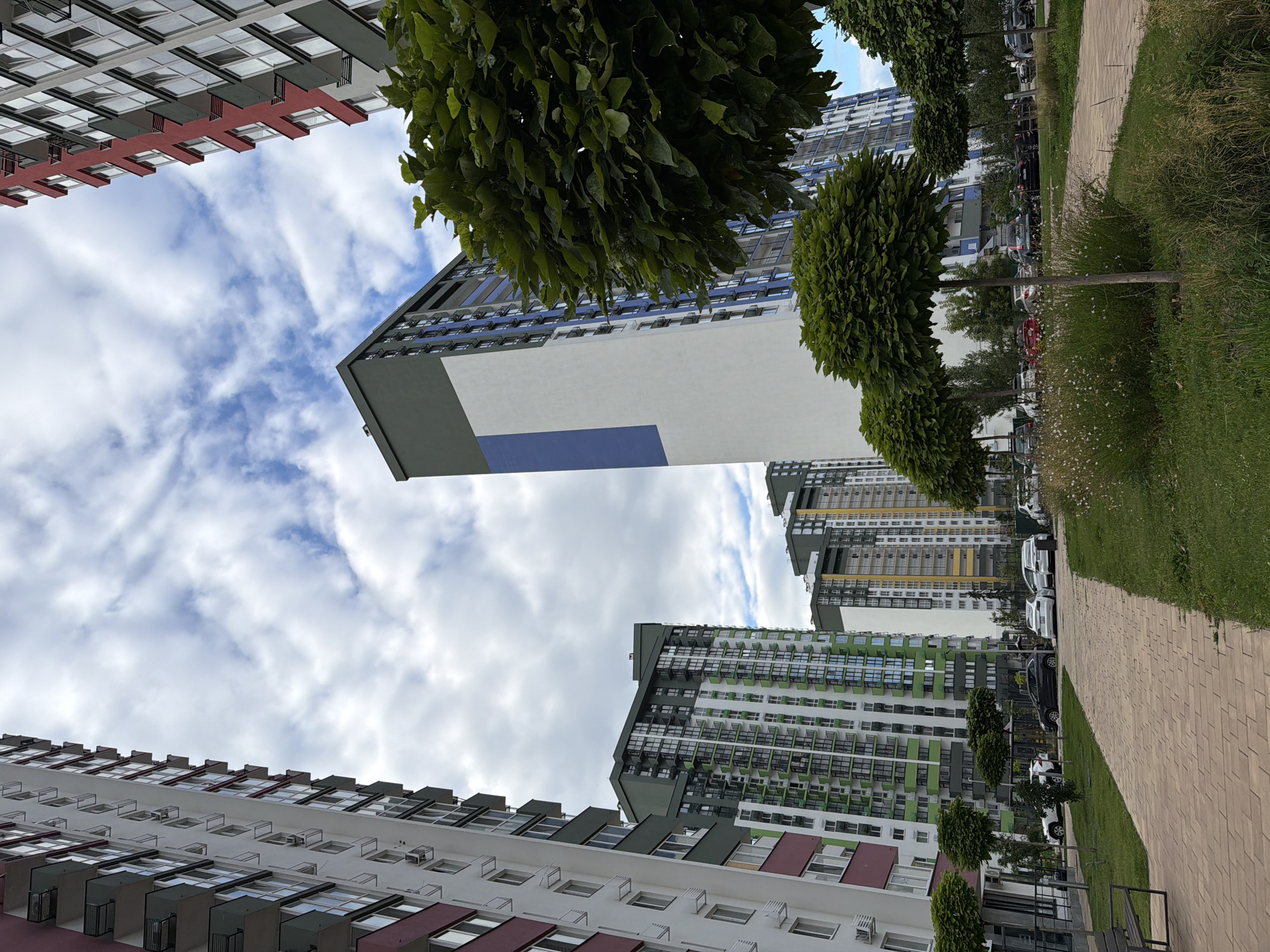 The image shows a modern residential complex in Kyiv, Ukraine, under a cloudy sky. In the foreground, a paved walkway lined with grass and ornamental trees leads towards several tall apartment buildings. The buildings are contemporary in design, with a mix of white, grey, green, and red facades, featuring numerous balconies and windows. Several cars are parked in a lot visible between the buildings and the walkway. The overall scene depicts an urban residential area with manicured landscaping.