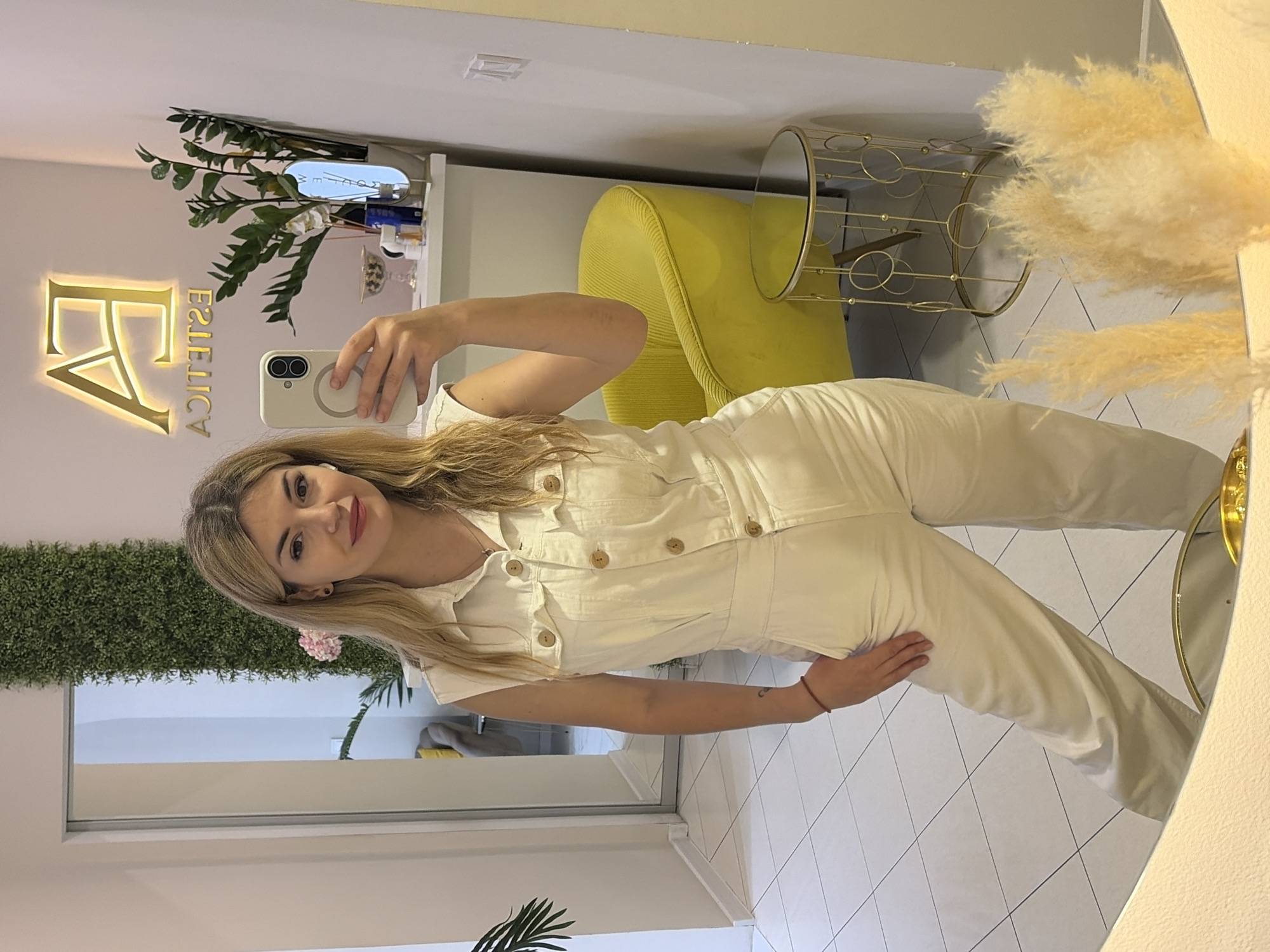 A young woman is taking a selfie in what appears to be the interior of a business named "Estetica," as indicated by a gold-toned sign behind her. The woman has blonde hair and is wearing a white jumpsuit with buttons down the front. She is holding a smartphone in her left hand. The background includes a yellow chair, a gold-toned table or stand with a mirror, and a wall with decorative plants. A window with more decorative greenery is visible on the left. The environment suggests a clean, modern aesthetic. The city, Budva, and country, Montenegro, are inferred only from the location name "Estetica Budva", which is assumed from the sign "Estetica" behind her in the image.