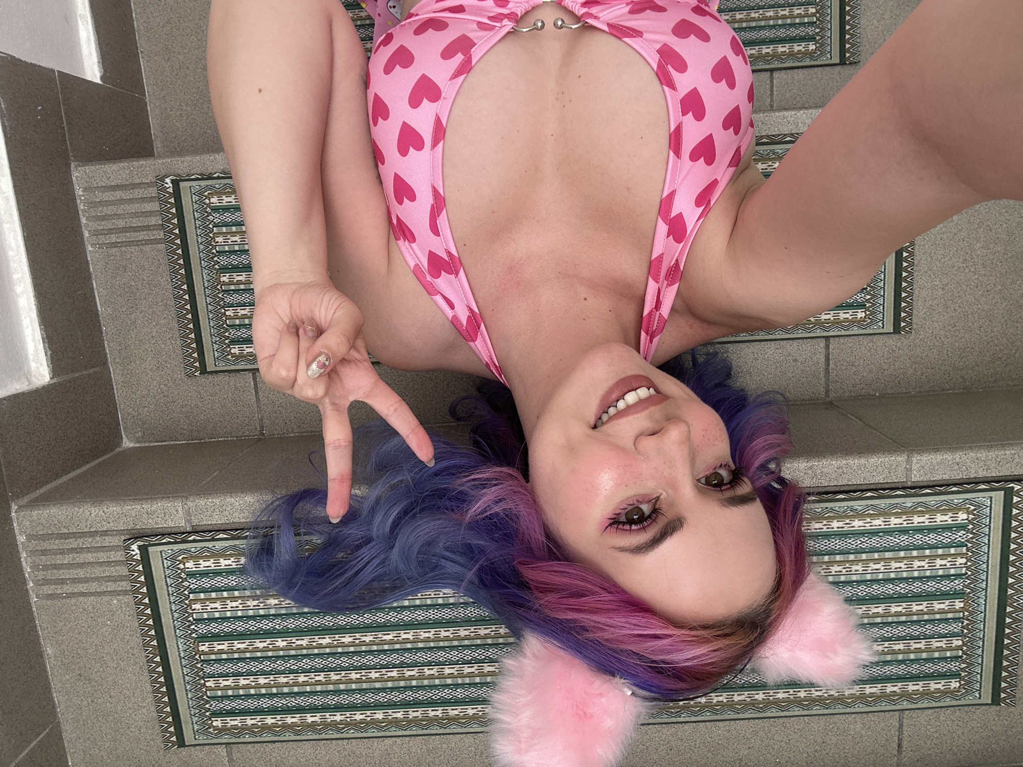 A woman is lying on a set of stairs, possibly in a building in Zagreb, Croatia. She is wearing a pink bathing suit with hearts printed on it and pink cat ears. Her hair is dyed blue and pink. She is making a peace sign with her left hand. The stairs are covered with small, rectangular patterned rugs.