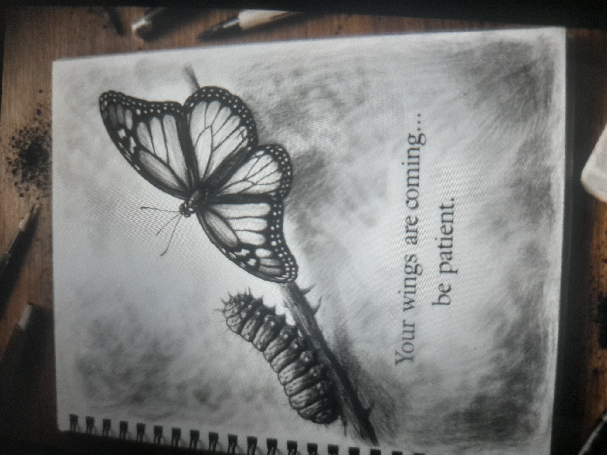 The image is a photograph of a sketchbook drawing. The drawing depicts a caterpillar on a thorny branch and a butterfly above it, both rendered in grayscale. Text beside the drawing reads "Your wings are coming... be patient." The sketchbook is bound with a spiral coil, visible along the bottom edge. The sketchbook rests on a wooden surface, with art supplies (pencil, charcoal, etc.) visible in the background around the edges of the sketchbook. No information in the image suggests the location is Akwanga, Nigeria.