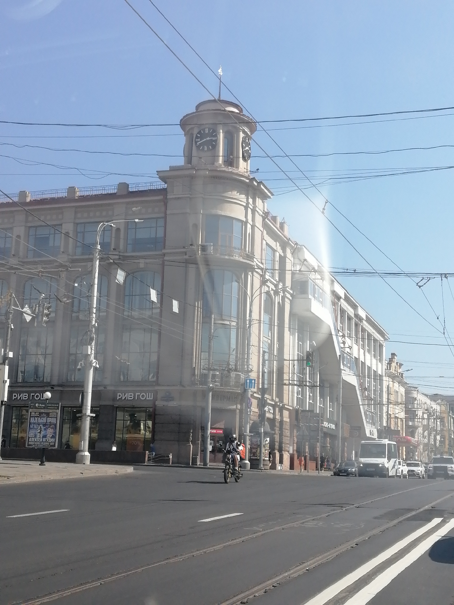 This image captures a street view in Rostov-on-Don, Russia. The dominant structure is a large, ornate building with a prominent clock tower. The building features numerous large windows, balconies, and intricate architectural details. Several signs with Russian text are visible, including "РИВ ГОШ". Overhead, a complex network of power lines and trolleybus wires crisscrosses the sky. In the foreground, a street with tram tracks runs across the scene. A person on a motorcycle is moving away from the viewer on the street. Several vehicles, including a white van, a white car, and a black car, are also present on the road. The sky is clear and blue, indicating a sunny day. The overall scene depicts an urban environment with a mix of historical architecture and modern traffic.