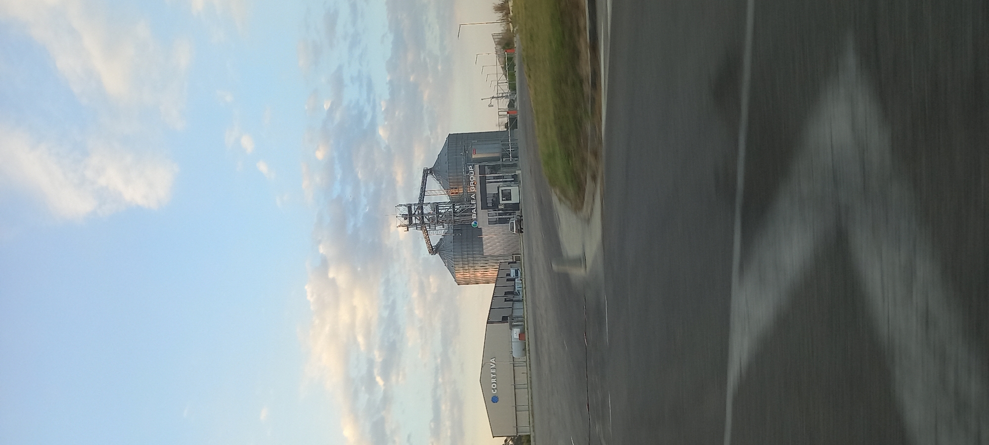A wide, low-angle, outdoor, vertical shot captured from a moving vehicle shows a large industrial facility with silos and buildings under a sky with scattered clouds. In the center of the frame, a complex of tall, cylindrical gray silos is visible, interconnected by a network of metal structures and chutes. The word "BALEA GROUP" is displayed in white text on one of the silos. To the left of the silos, a long, rectangular building with the word "CORTEVA" in blue letters is situated. A small white security booth is positioned in front of the entrance to the BALEA GROUP facility. A single silver car is parked near the security booth. The foreground of the image is dominated by a gray asphalt road with white lane markings. The background consists of a grassy area and some distant trees. The overall impression is one of an agricultural processing or storage facility located on the outskirts of an urban area. The image was taken in the vicinity of Dobrich, Bulgaria.
