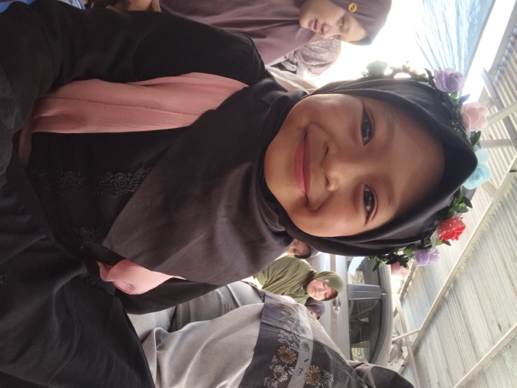 A young girl wearing a black hijab adorned with a floral crown is the central subject of the image, smiling directly at the camera. She is dressed in a black garment with a pink accent. In the background, other individuals are visible. One woman to the left wears a purple hijab, and another to the right wears a green hijab. Further in the background, a vehicle is parked. The setting appears to be outdoors, under a corrugated metal roof structure, possibly in the city of Kepuh Kiriman, Indonesia.