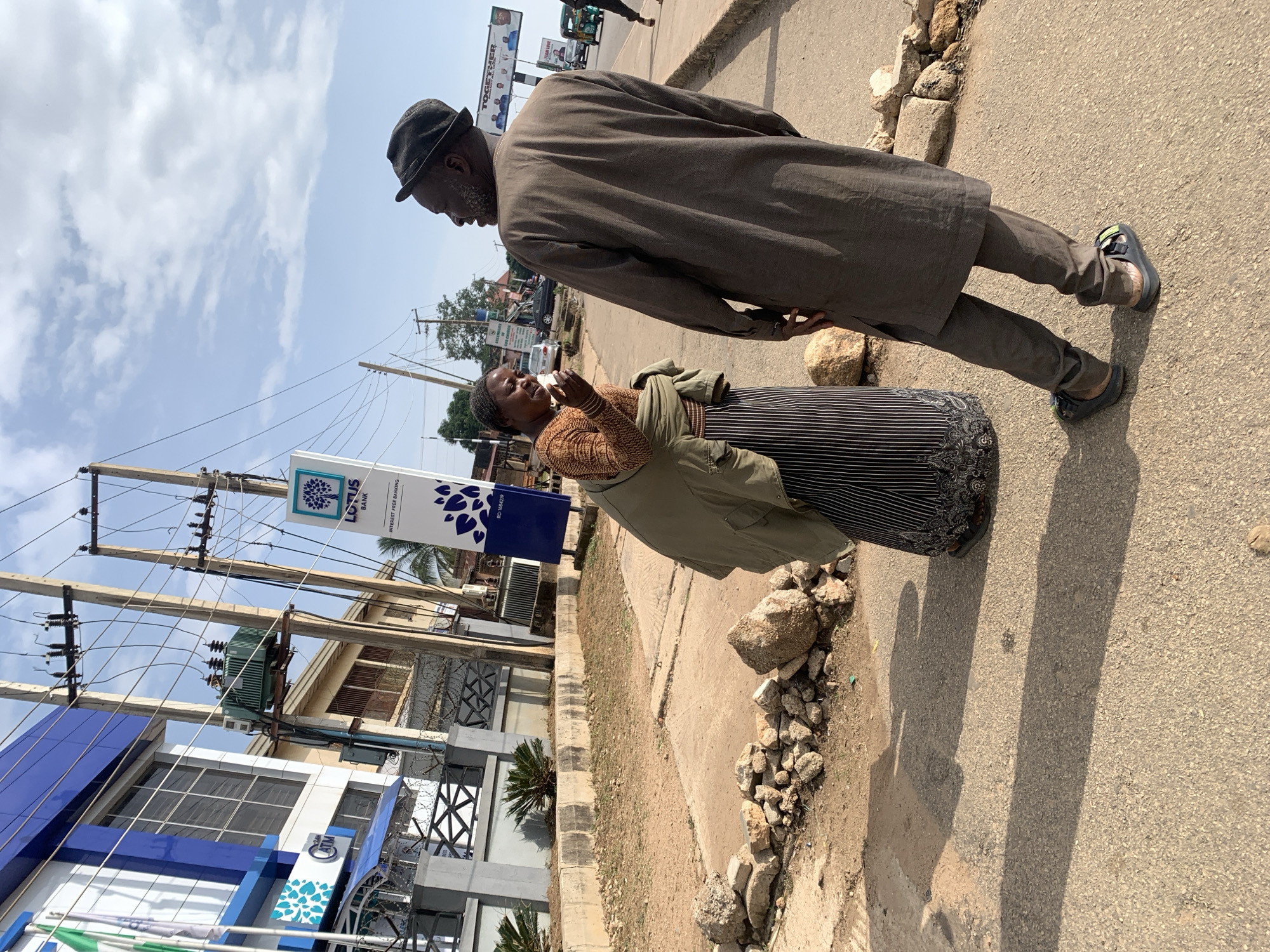 The image depicts an outdoor scene in Jos, Nigeria, with two individuals interacting on a paved street. A woman, wearing a brown, patterned long-sleeved shirt and a dark, vertically striped skirt with a black trim at the hem, stands facing a man. She is holding a white object, possibly a cup or a tissue, to her mouth. A green jacket is draped over her shoulders.
The man, wearing a dark grey, long-sleeved tunic and matching trousers, is facing away from the camera, looking towards the woman. He has a dark-colored hat on his head and is wearing dark sandals with yellow and green accents.
In the background, a modern building with blue and white accents is visible, with the "LOTUS BANK" logo displayed on a sign. Power lines and a transformer are also prominent in the background, as is a small, green transformer mounted on a pole. The street itself is bordered by a concrete barrier and a dirt area with scattered rocks and debris. Further in the background, other buildings, vehicles, and street signs are visible, indicating an urban environment. The sky is partly cloudy. The overall impression is a typical street scene in a Nigerian city.
