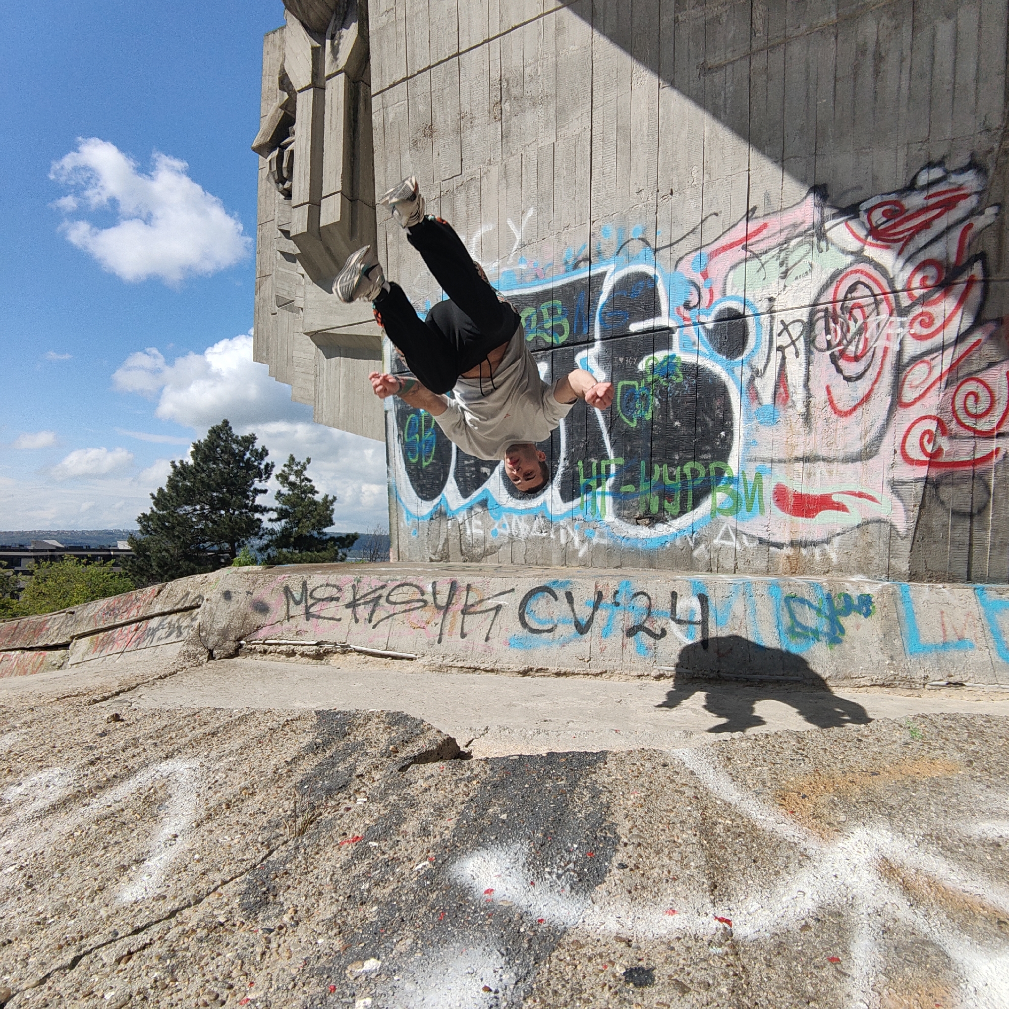 A man is performing a backflip in front of a concrete monument covered in graffiti. The location is in an urban area, with trees and buildings visible in the background under a blue sky with clouds. The man is wearing a light-colored shirt and dark pants, with sneakers. His shadow is cast on the ground in front of him. The graffiti on the monument appears to be multicolored, with various tags and designs.