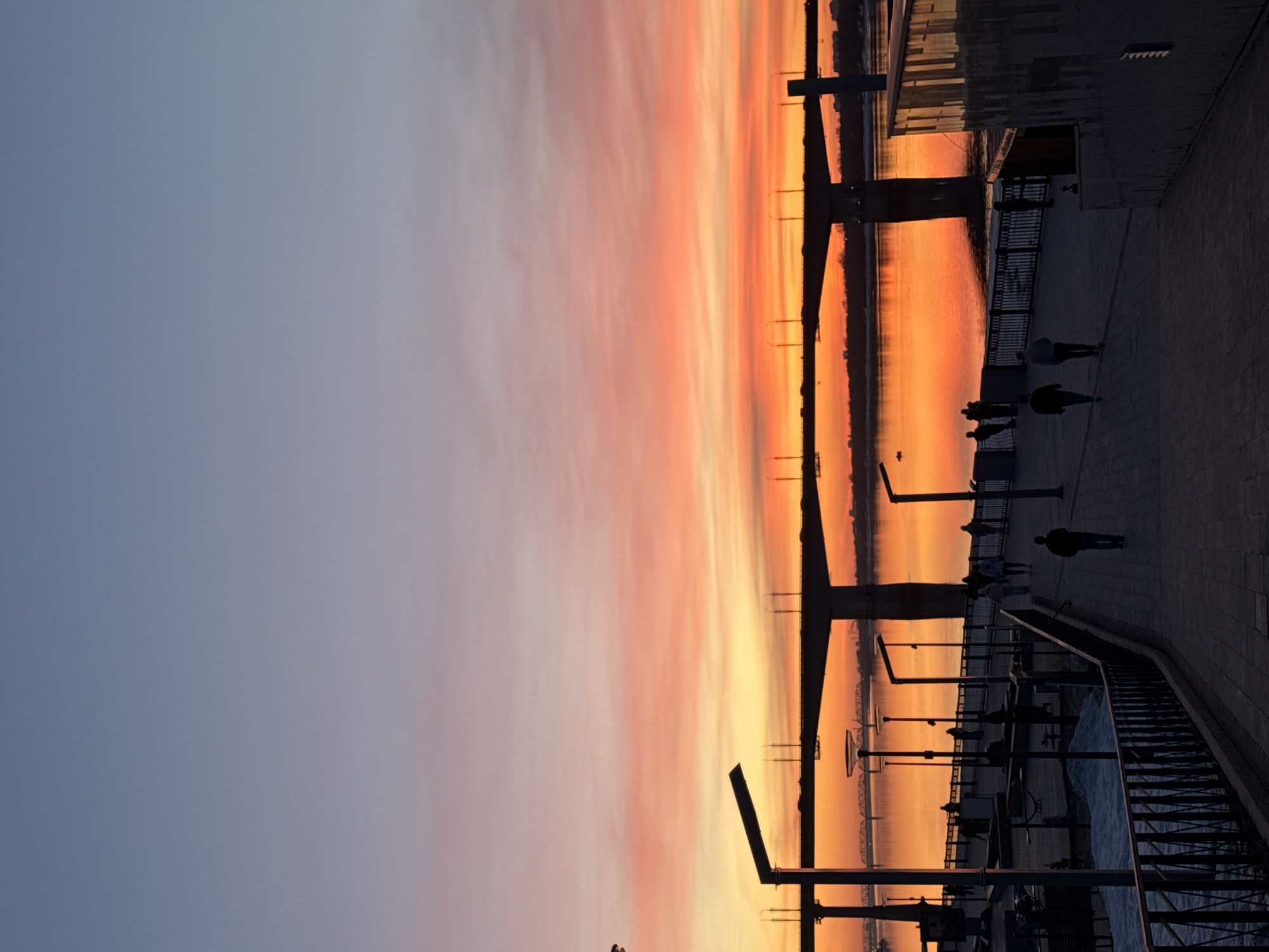 The image shows a sunset scene in what appears to be Perm, Russia, specifically on a riverfront walkway. The sky is a gradient of colors, transitioning from a soft blue-gray to vibrant shades of pink, orange, and yellow near the horizon. The sun's reflection is prominently visible on the water's surface, creating a shimmering, orange glow.
A walkway runs along the river, featuring metal railings and streetlights with unique, elongated lamps. Silhouetted figures of people are visible strolling along the walkway, indicating an evening ambiance. The silhouettes include individuals and small groups.
In the background, the silhouette of a bridge stretches across the river. The overall atmosphere is tranquil and scenic. The perspective is slightly tilted.