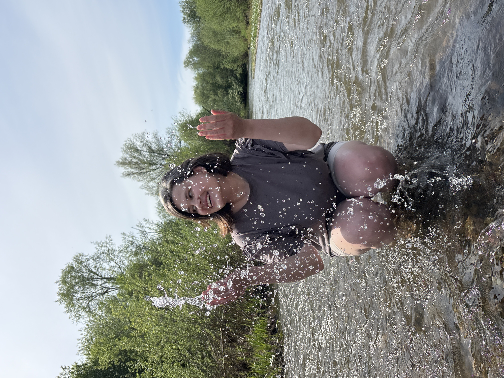 A woman is squatting in a river, splashing water with her hands. The background consists of trees and the sky. The location is Усть-Тунтор, Russia.