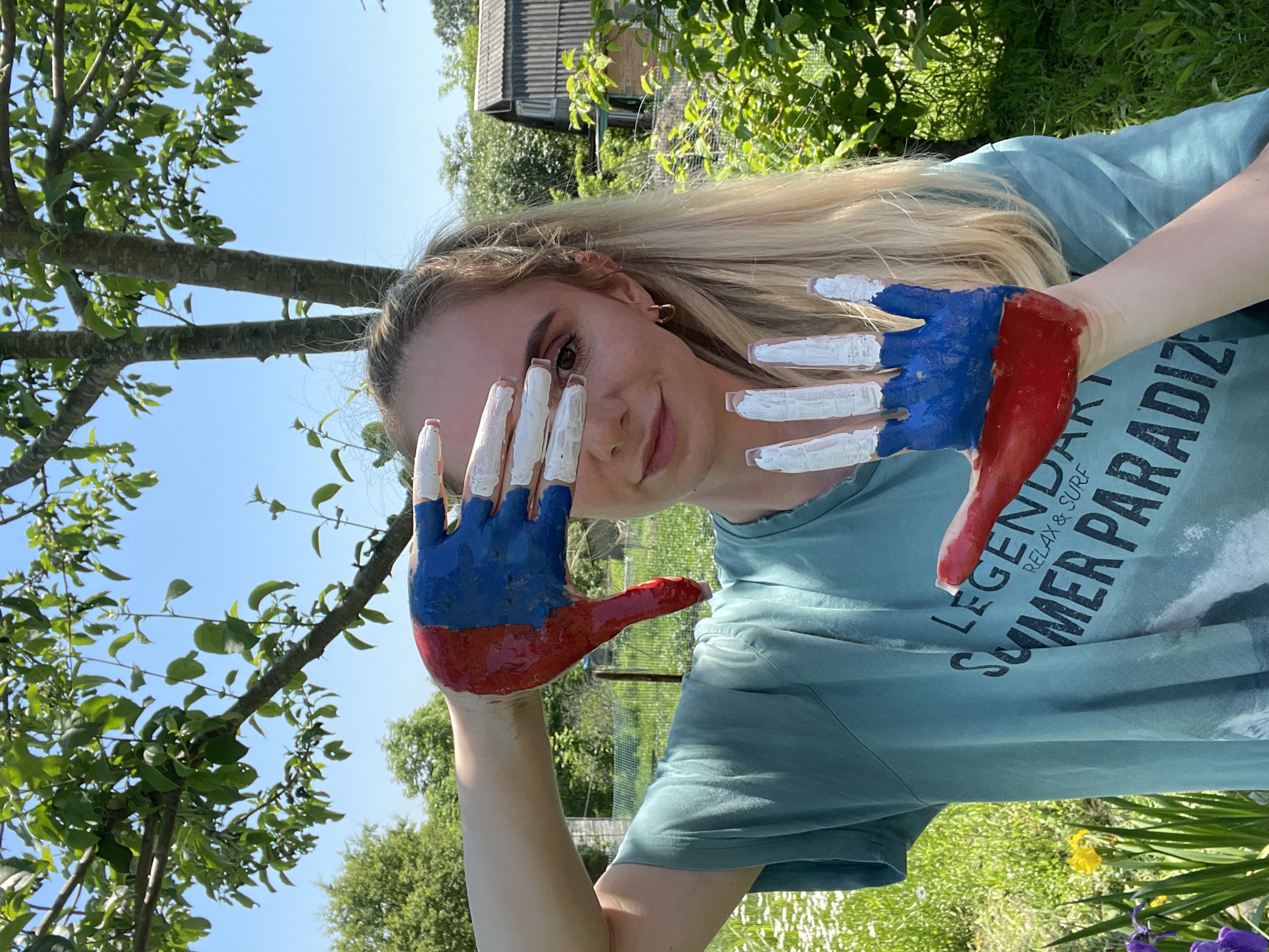 A young Caucasian woman with blonde hair pulled into a ponytail stands outdoors, partially obscuring her face with her hands, which are painted with red, white, and blue. She is wearing a teal t-shirt with the words "LEGENDARY SUMMER PARADIZE RELAX & SURF" printed on it. Her right hand, painted in red and blue, is held up to her face. Her left hand, painted white on the fingers and blue and red on the palm, is held in front of her. The background shows lush green trees, foliage, a wooden fence, and a rustic wooden building, suggesting a rural or suburban setting. The sky is clear and blue. The overall lighting indicates daytime. This scene is likely located in the city of Yasnoe, Russia, given the context of the provided information.