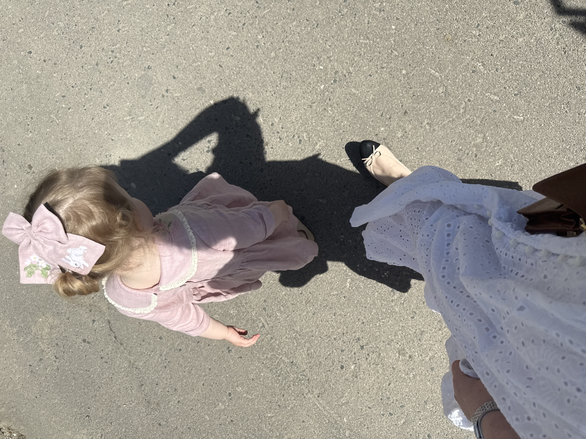 A high-angle shot shows a child and an adult on a gray concrete surface. The child, wearing a pink dress with lace trim and a pink hair bow with an embroidered rabbit, is positioned on the left. The adult, wearing a white eyelet dress and black and beige flats, stands on the right. A dark shadow is cast over the scene, partially obscuring the child. The photo might be taken in Penza, Russia, but there are no elements to confirm it.