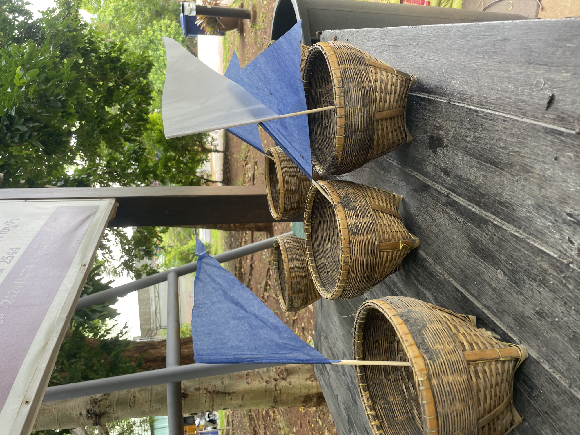 The image shows four woven bamboo baskets, each with a flag attached by a wooden stick, arranged on a dark wooden surface. Three of the baskets have blue flags, and one has a white flag. The baskets appear to be empty. In the background, there are trees with green foliage, a metal support structure, and a partially visible sign with writing. The scene suggests an outdoor setting, possibly in a park or near a natural area. The general environment is that of Na Chom Thian, Thailand, with tropical vegetation and a casual outdoor setup.