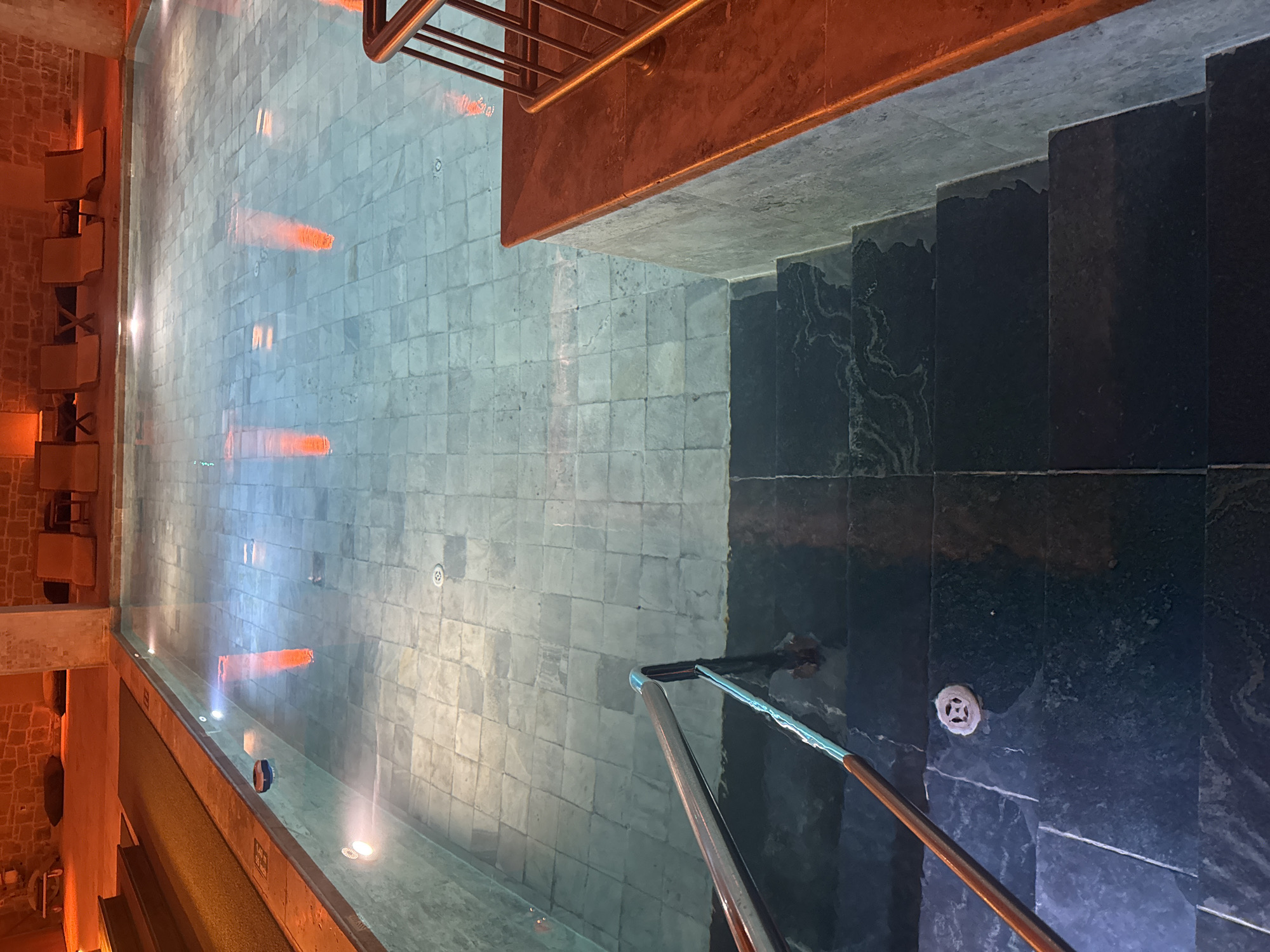 The image shows an indoor swimming pool with glass walls, likely located in a hotel or spa in Ürgüp, Turkey, as the architecture and lighting suggest a luxury environment commonly found in the region's cave hotels.
The pool itself has a tiled bottom with embedded lights creating reflections. Adjacent to the pool are dark-colored stone stairs leading downwards. A metal railing is visible alongside the stairs and at the edge of the pool. A series of chairs or seating is located on an elevated ledge along one side of the pool, illuminated with warm lighting. The pool's water is still, mirroring the surrounding environment.