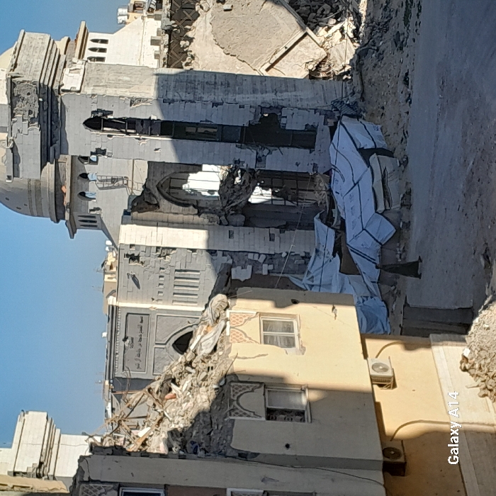 A daytime photograph captures a scene of extensive structural damage in Khan Yunis, Palestinian Territory. In the midground, the Great Mosque of the City (الجامع الكبير للمدينة), identified by Arabic script on its facade, stands heavily damaged. Its central minaret or tower features a dome, and significant portions of its masonry walls have collapsed, revealing interior elements. Adjacent structures of the mosque complex are similarly compromised. In the foreground left, a multi-story building is partially collapsed, with rubble on its upper section and intact windows with decorative trim visible on lower floors. An exterior air conditioning unit is mounted on its wall. The unpaved ground surface is covered with debris and scattered piles of rubble. Several white tarpaulins or sheets are visible on the ground between the foreground building and the mosque. A lone individual, clad in dark attire, walks on the dirt path, moving away from the viewer. The sky is clear and blue.