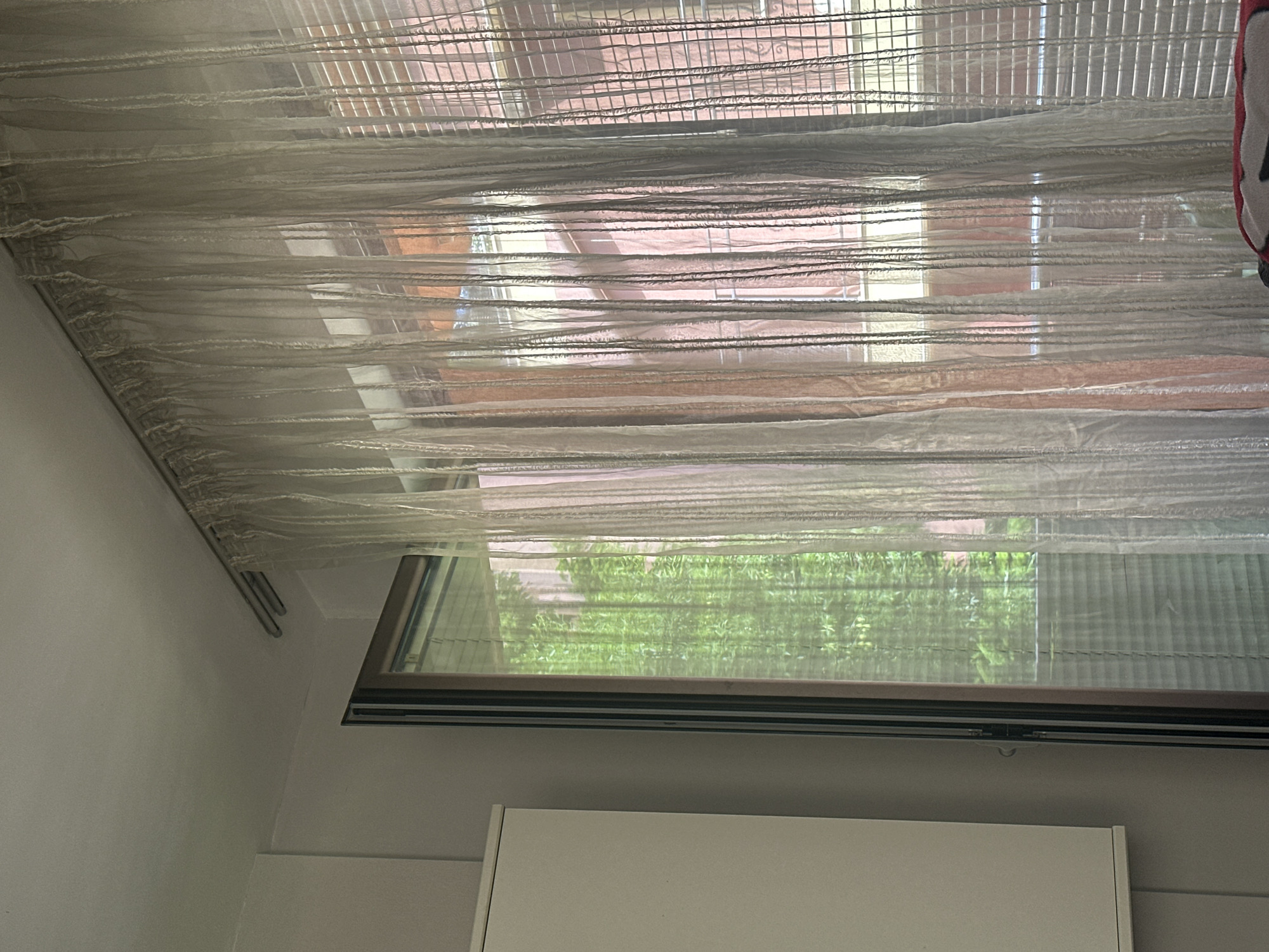The image shows a window with sheer, horizontal blinds in a room. Outside the window, greenery suggests trees or a garden. The room has a white wall and a white cabinet below the window. The exact location in Sofia, Bulgaria, cannot be determined without further information. There are no people in the image, no actions depicted.