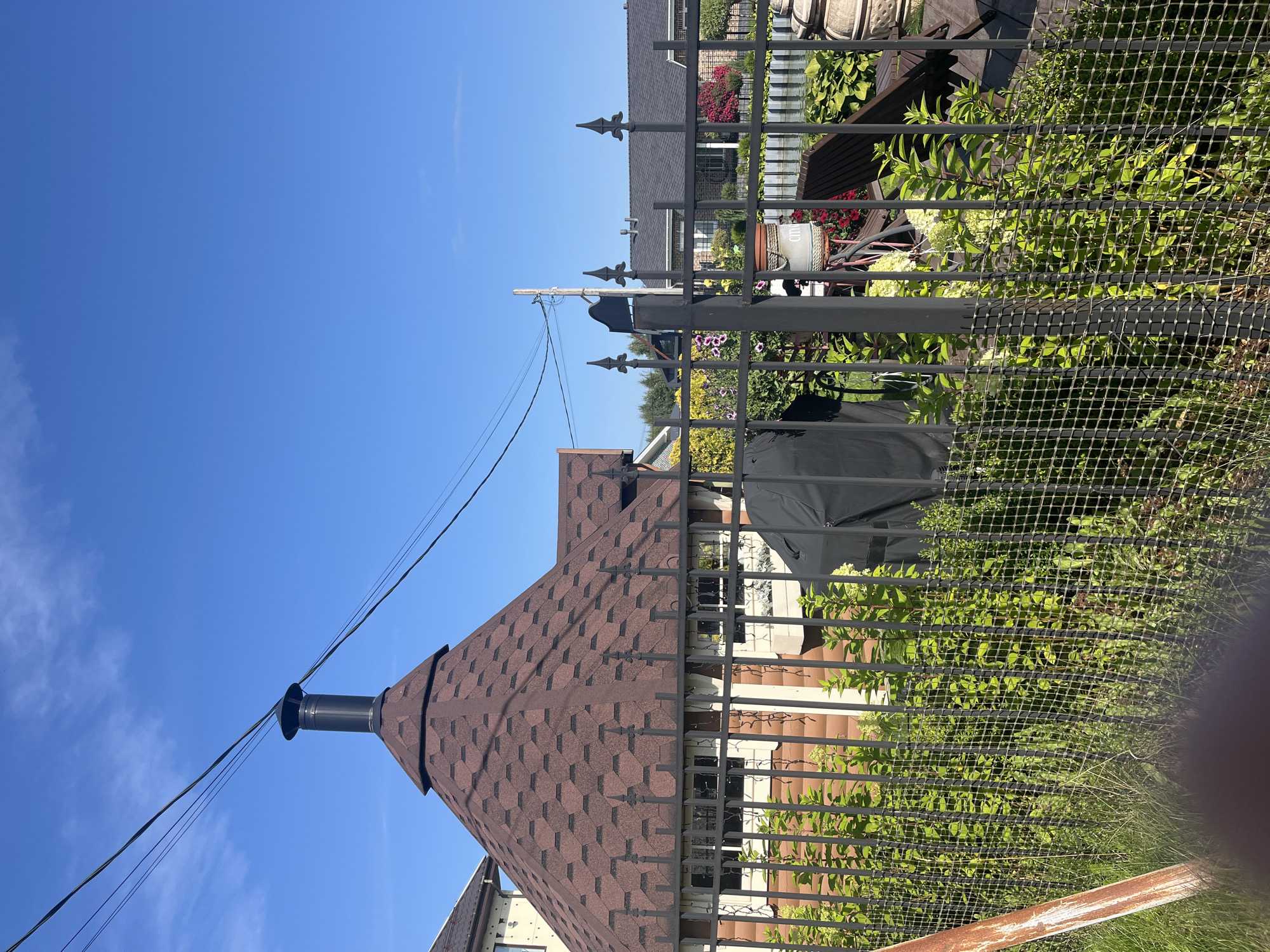 In the city of Babigonskaya Valley, Russia, a clear blue sky dominates the upper portion of the image. A conical, brown-shingled roof with a black chimney extends from a building on the left. Power lines cross diagonally through the frame. In the foreground, a black metal fence with a mesh backing encloses a yard. Beyond the fence, lush green foliage and various plants are visible. To the right of the conical roof building, another building with a dark gray roof is partially visible. The environment appears to be a residential area with well-maintained landscaping.